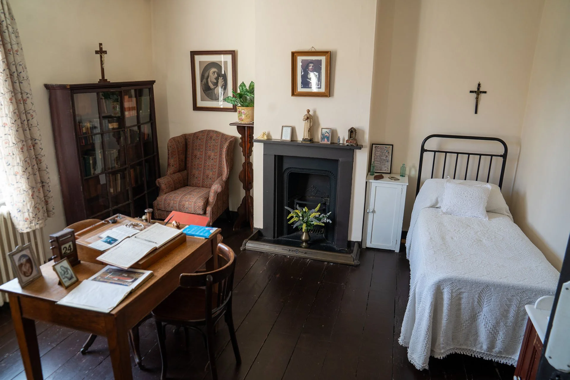 A cozy room with a single bed covered in a white lace bedspread, a black metal headboard, a small white cabinet next to the bed, a fireplace with religious figurines and framed photos on the mantel, a patterned armchair, a tall bookshelf filled with books, a wooden table with open books, framed photos, and a desk calendar, a window with floral curtains, and a crucifix on the wall.