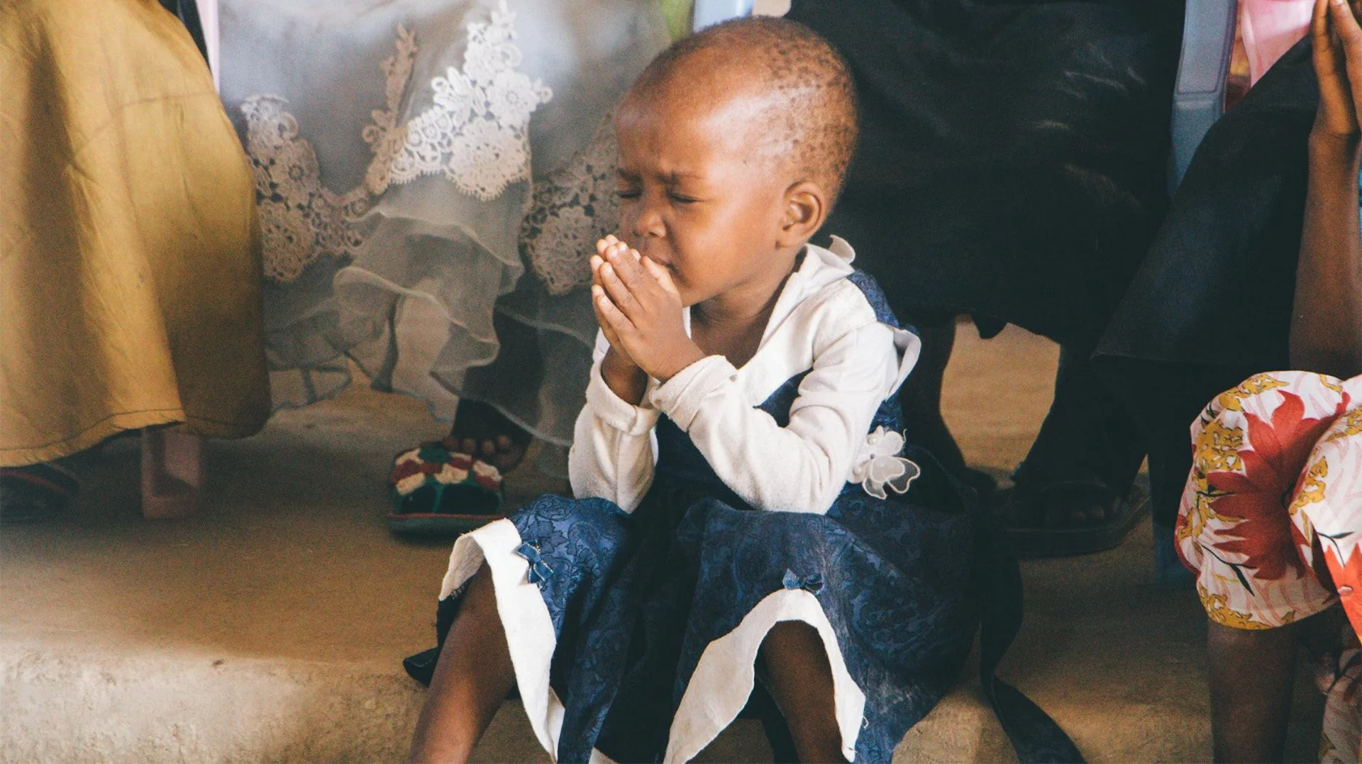 A young girl with short hair, sitting on the floor with her eyes closed and hands clasped together to pray.