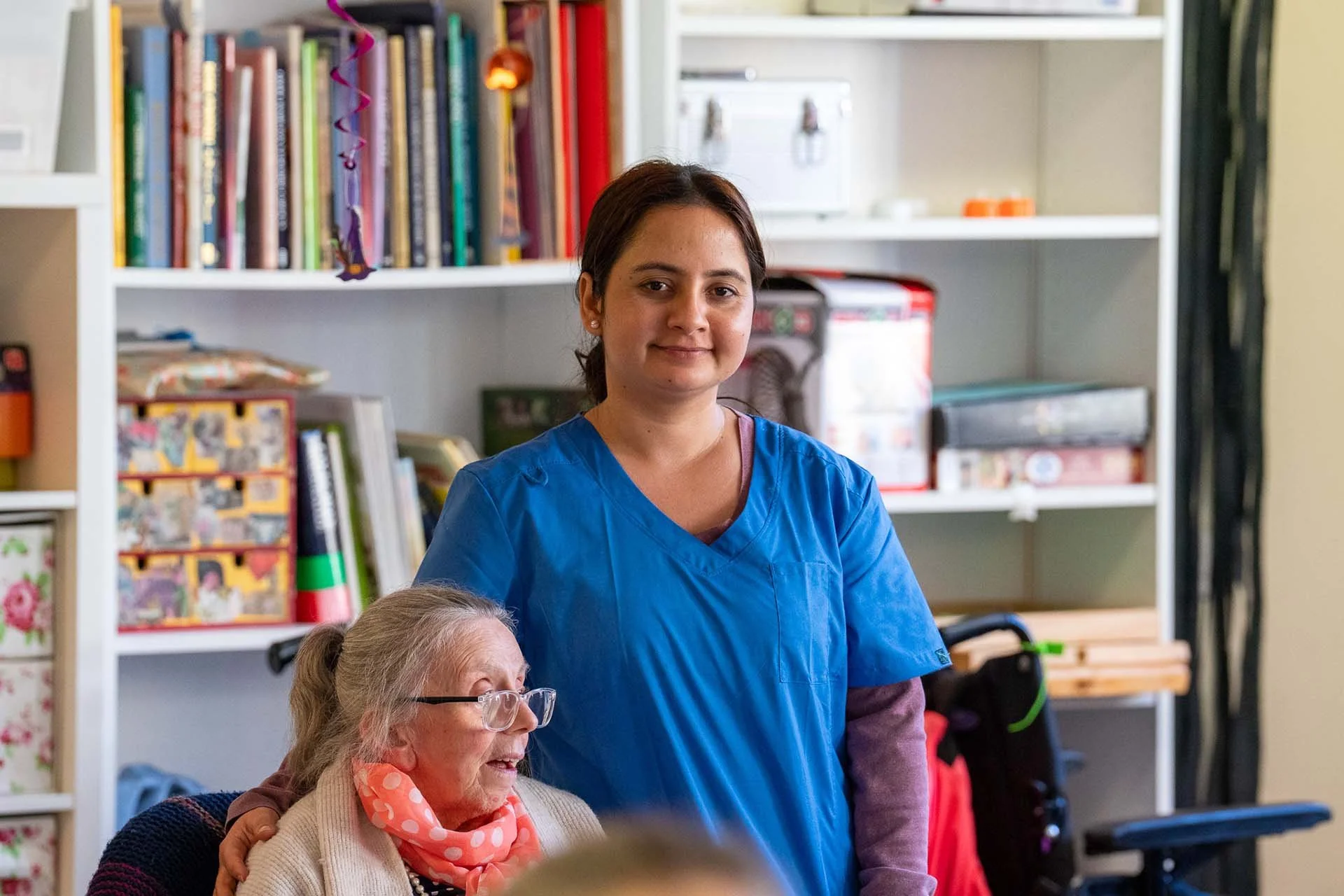 A young woman wearing a blue medical scrub standing with her hand on an elderly woman's shoulder in a bright room with bookshelves and medical equipment.