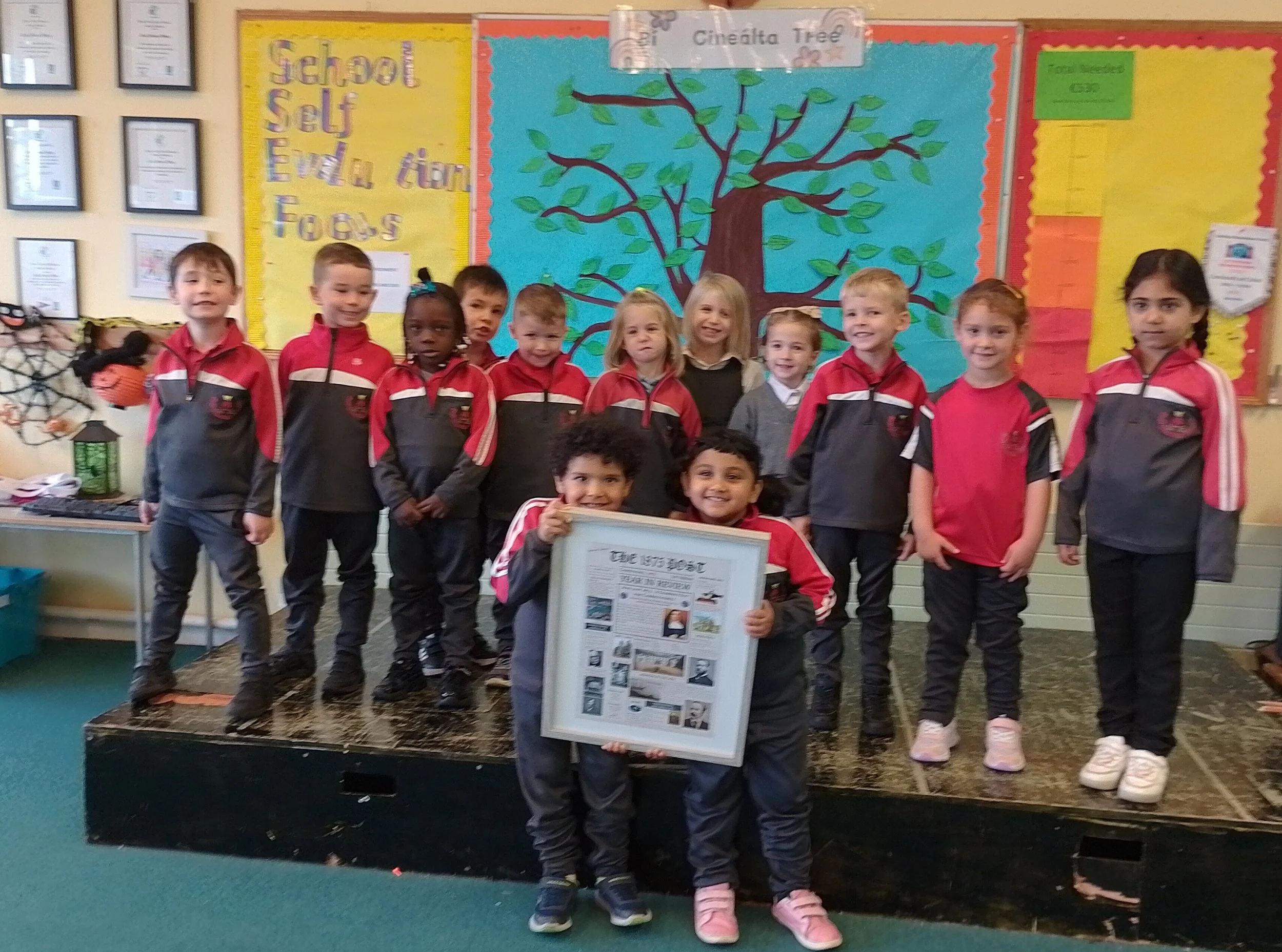 Group of young children in school uniforms standing on a stage, holding a framed newspaper titled "The Inver Post," with a colorful classroom bulletin board featuring a large paper tree behind them.