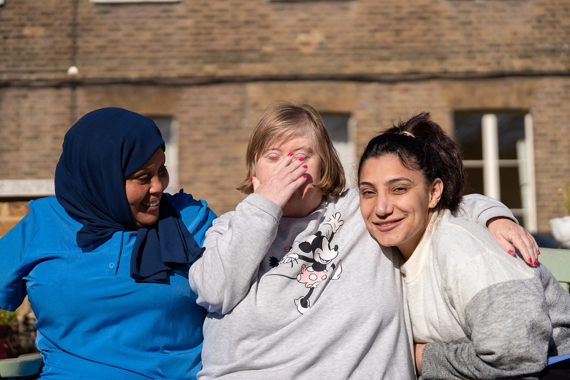 Three women sitting outside, smiling and embracing, one in a headscarf, one with hand covering face, and the third with dark multi-colored hair.