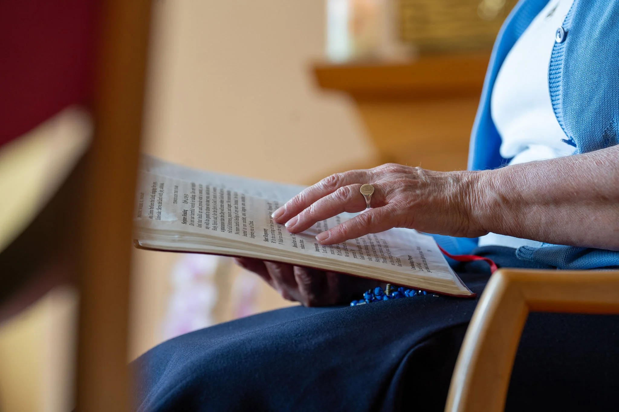 Close-up of an elderly person's hands reading a book, wearing a ring, sitting in a wooden chair.