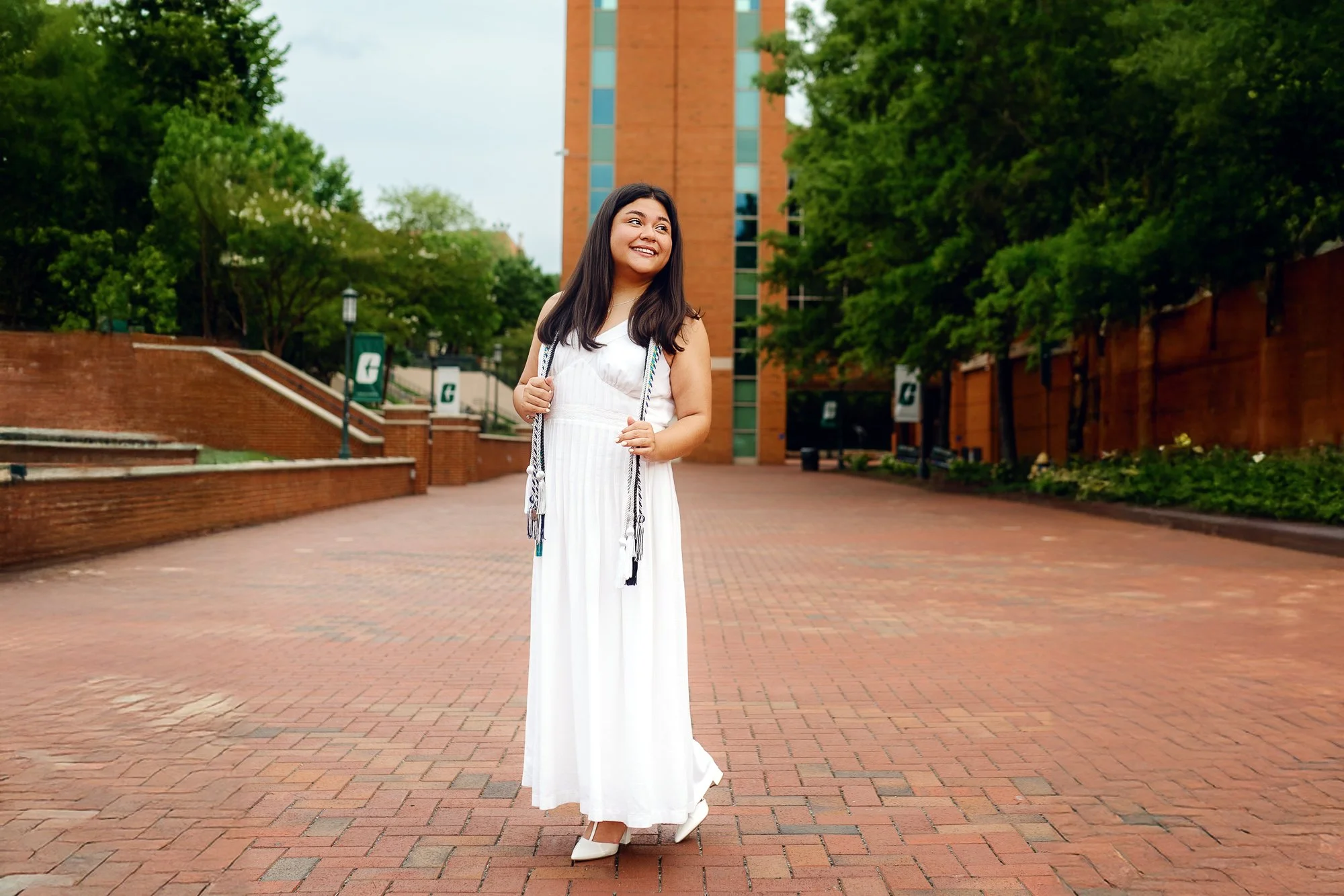 Ashley smiling in a white graduation dress while standing in the brick plaza near UNC Charlotte's Bell Tower