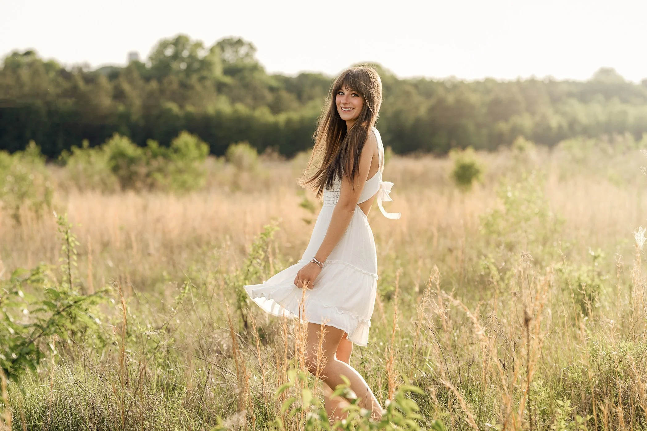 A young woman in a white dress walking through a grassy field with trees in the background during daylight in Harrisburg NC