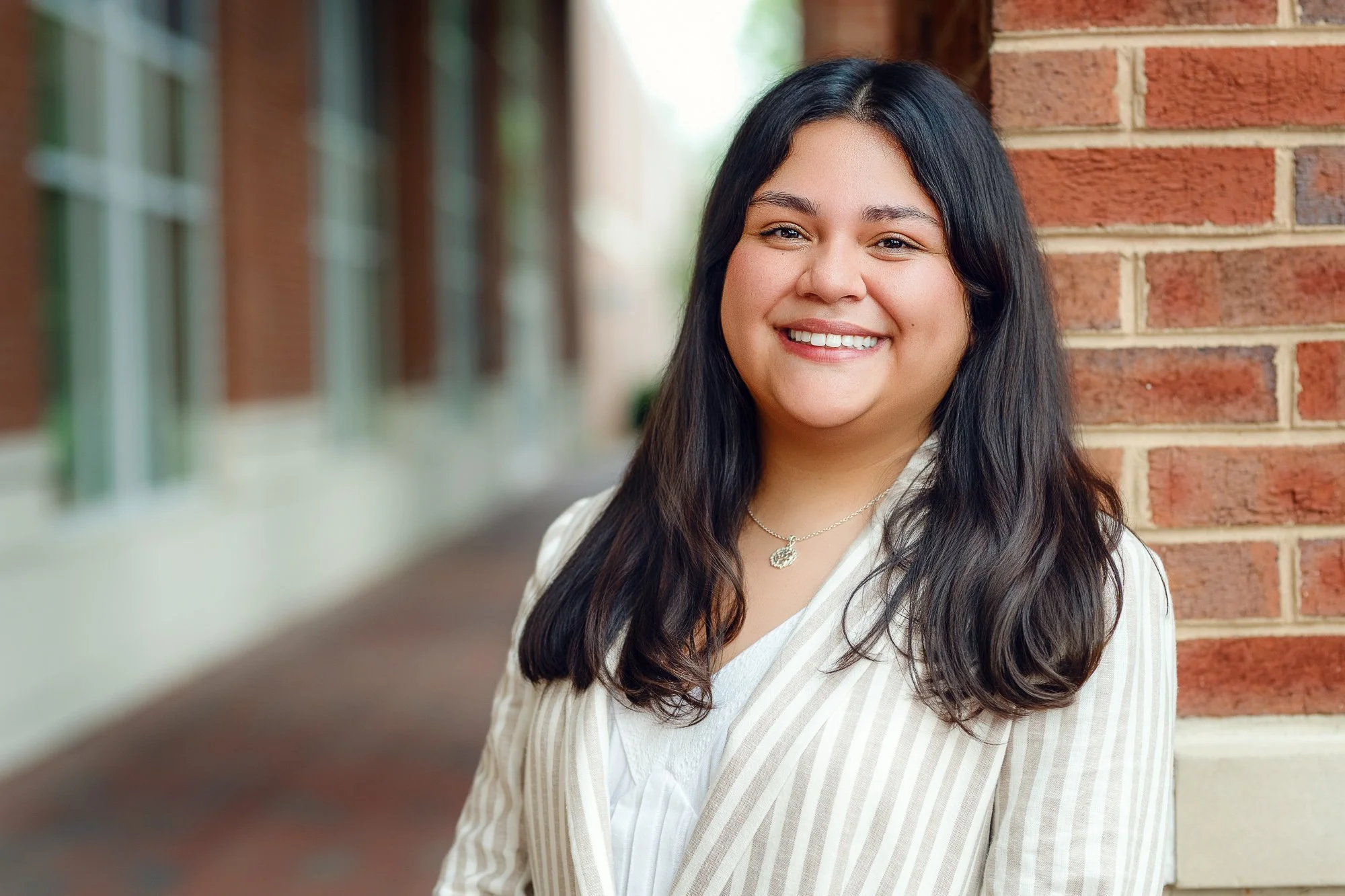 Portrait headshot of a smiling young woman with long dark hair, wearing a light-colored striped blazer and a necklace, standing outdoors near a brick wall.