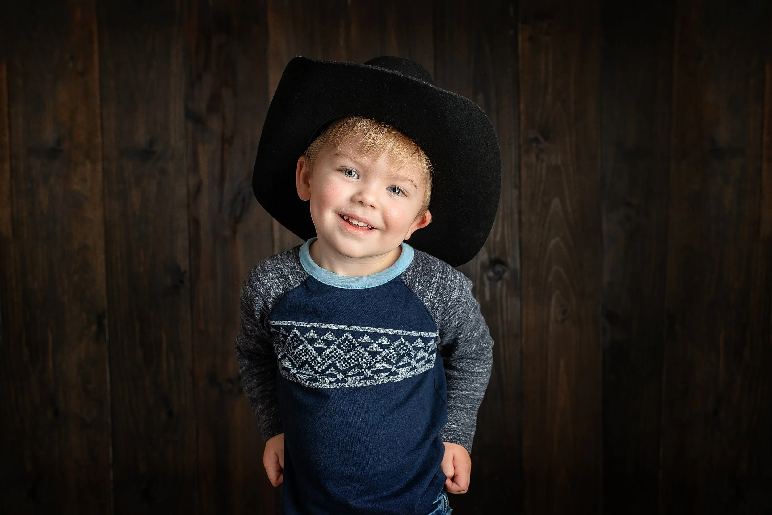 A young boy with blond hair and blue eyes wearing a black cowboy hat and a blue and gray sweater with a geometric pattern, smiling at the camera, standing against a dark wooden background.