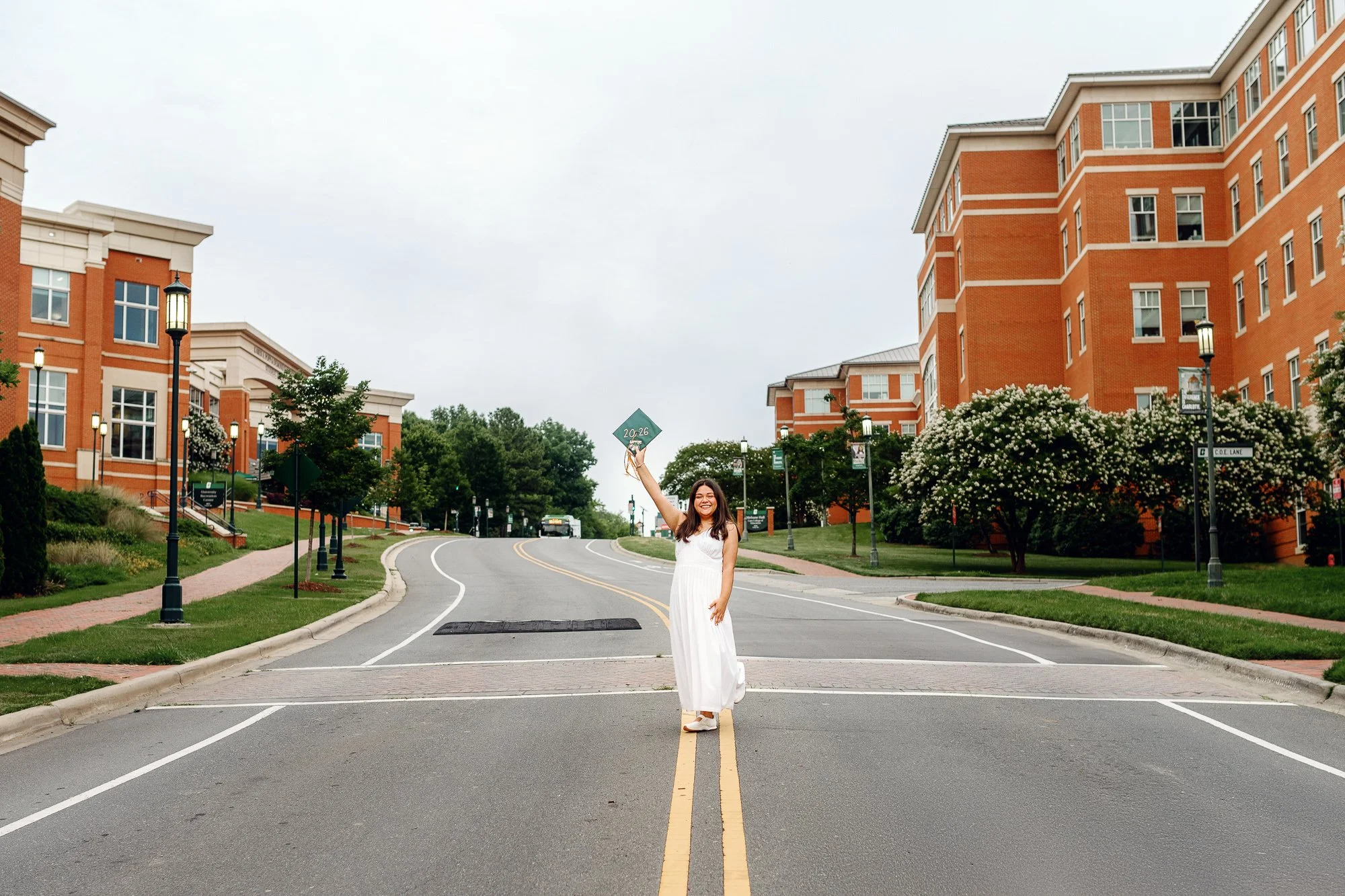 Ashley standing in the center of a campus roadway holding her graduation cap above her head.
