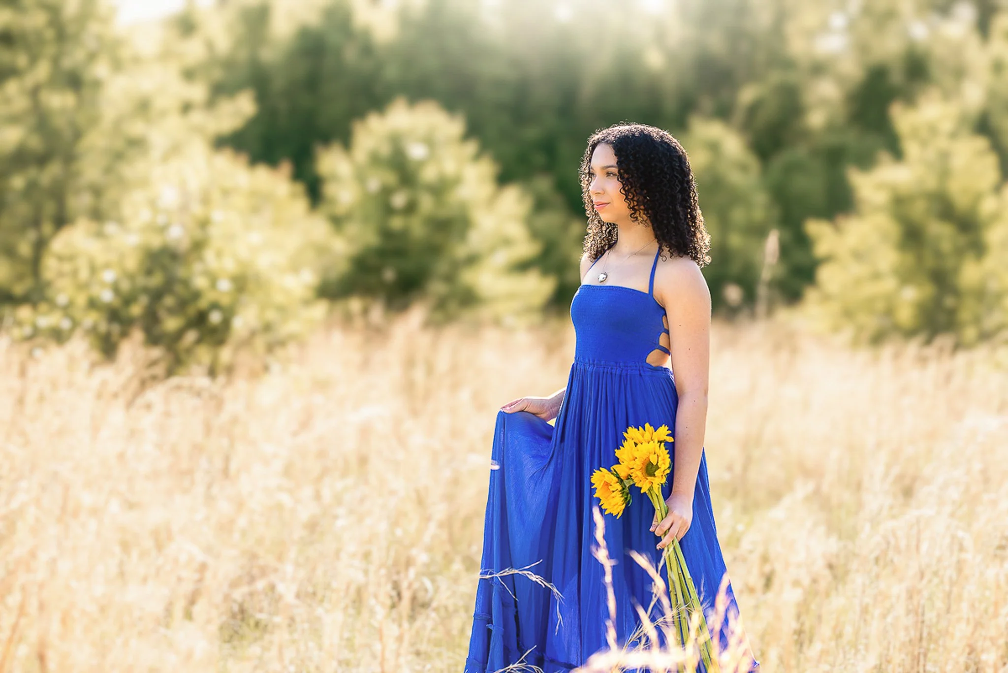 high school senior girl in blue dress in yellow green field holding sunflowers