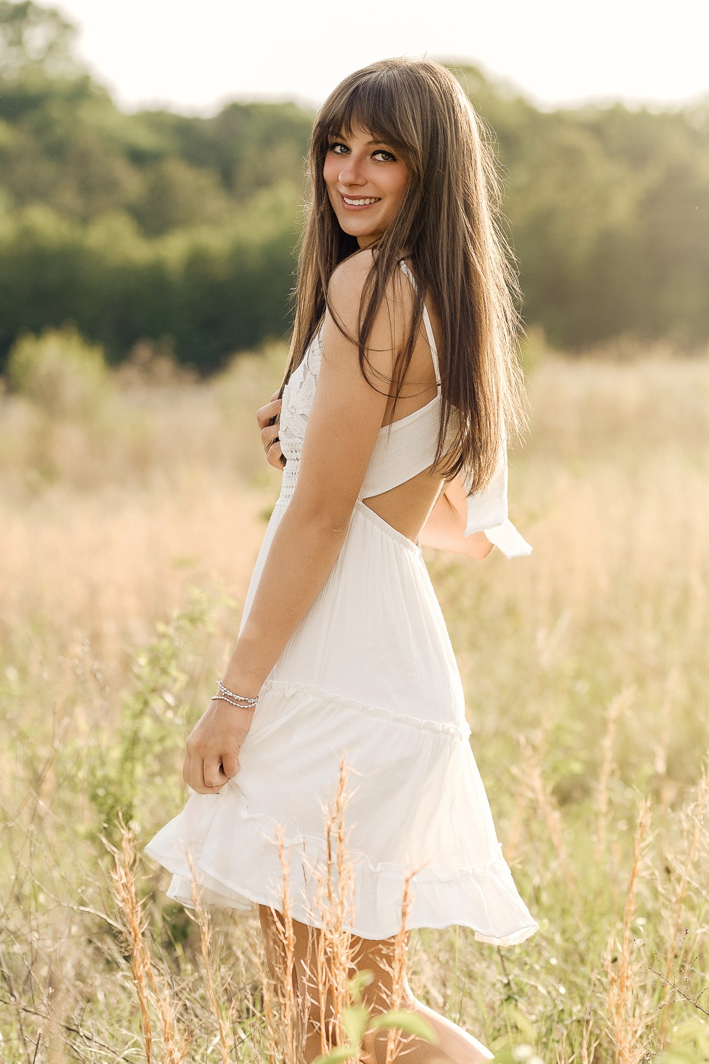 A young woman with long brown hair, wearing a white dress, smiling outdoors in a field with tall grass and trees in the background in Harrisburg NC
