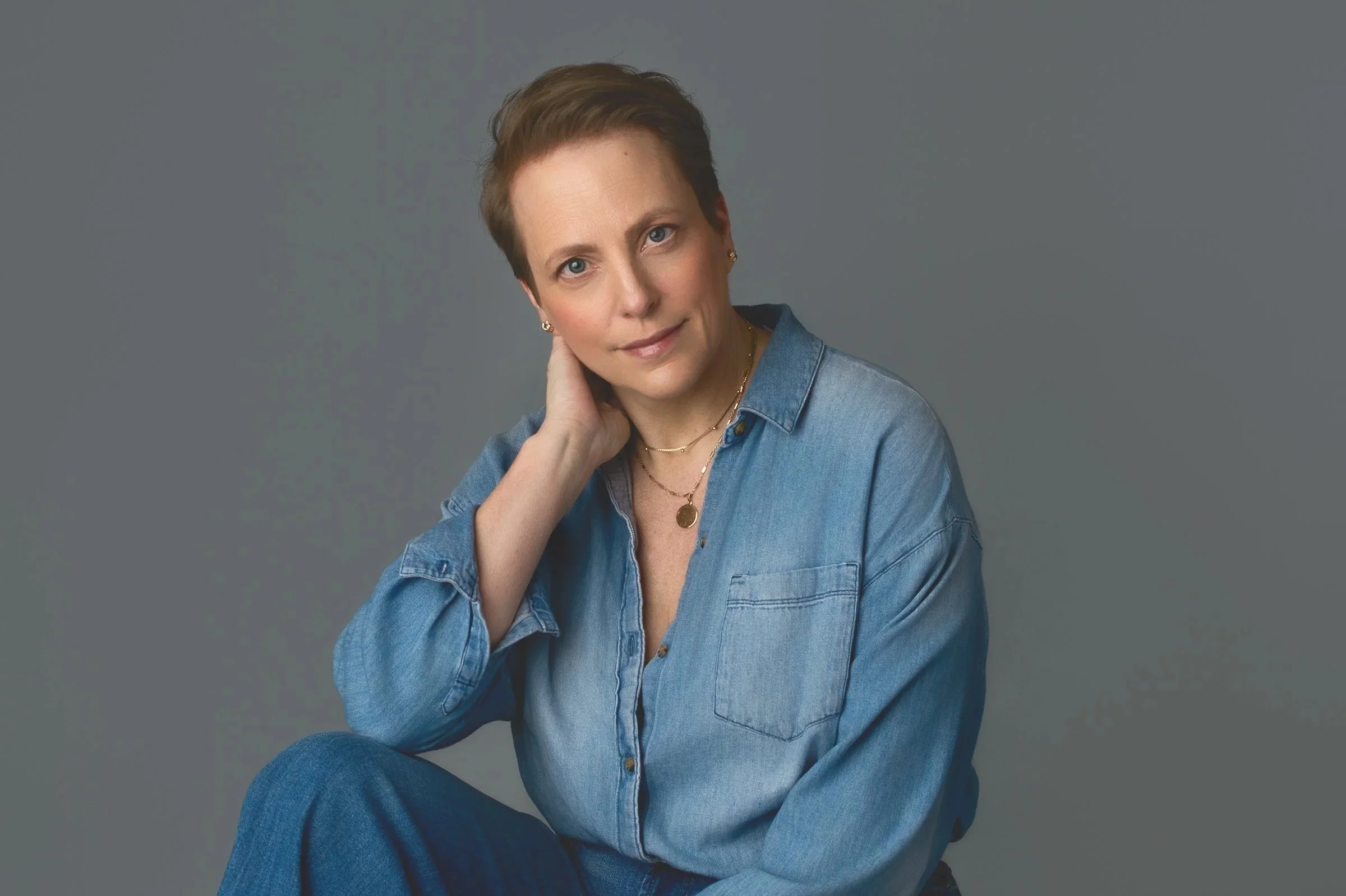 A woman with short brown hair wearing a denim shirt and gold jewelry posing against a grey background.