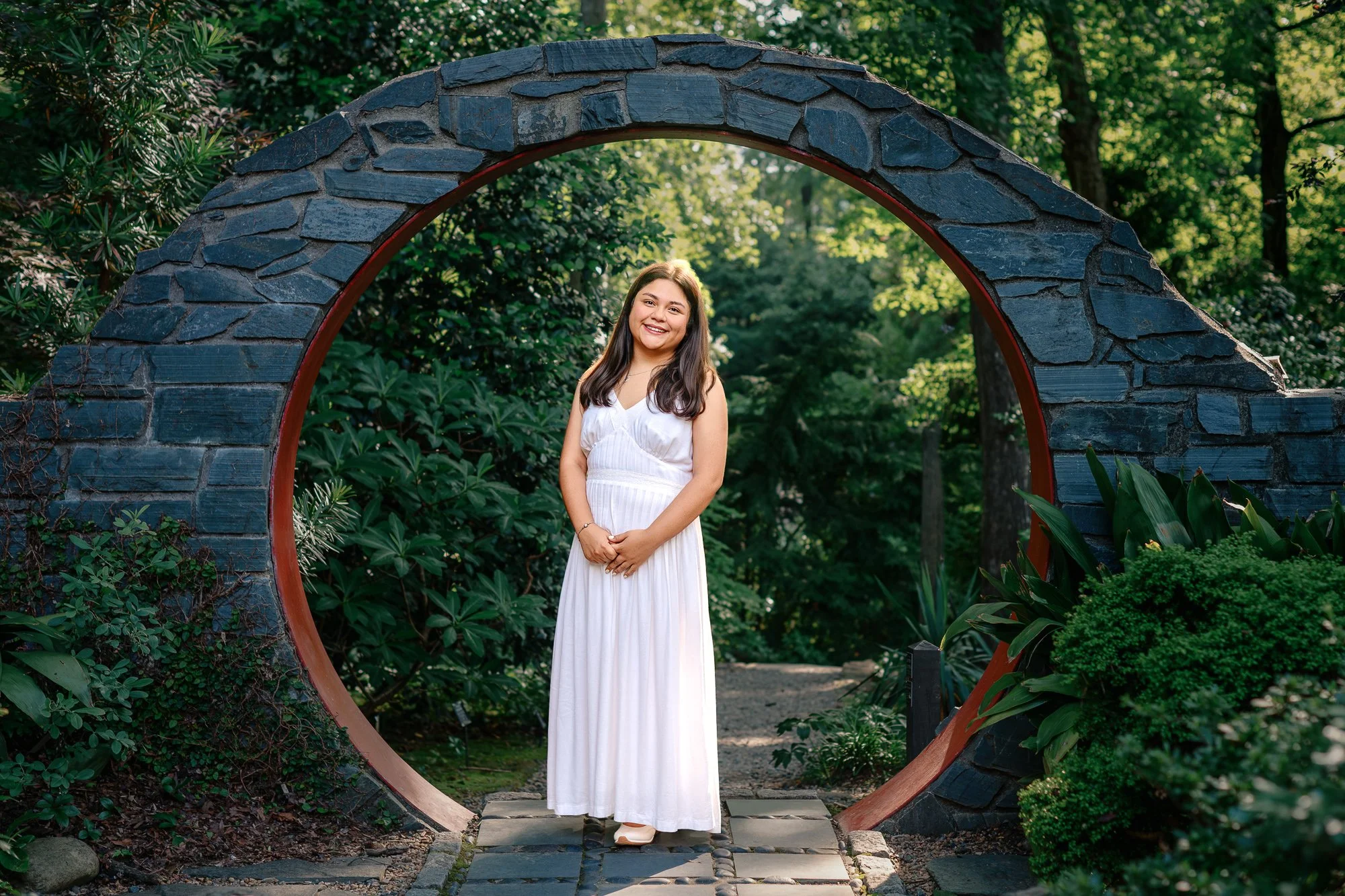 Ashley standing on a garden path in a flowing white dress surrounded by lush greenery at UNC Charlotte