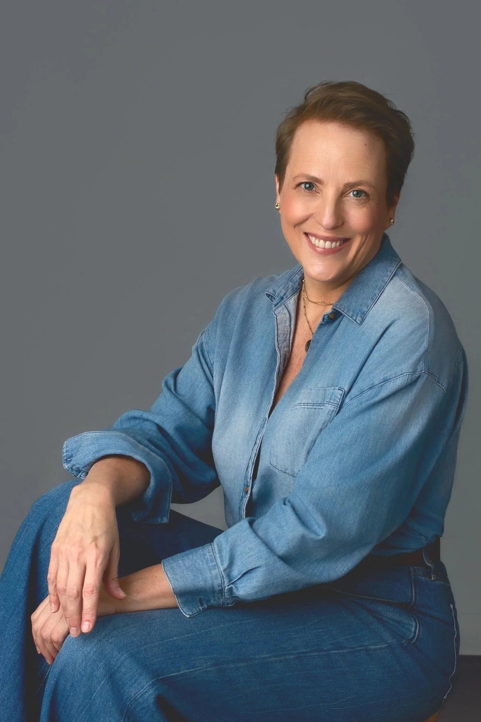 A woman with short brown hair, smiling, wearing a denim shirt and jeans, sitting against a gray background.