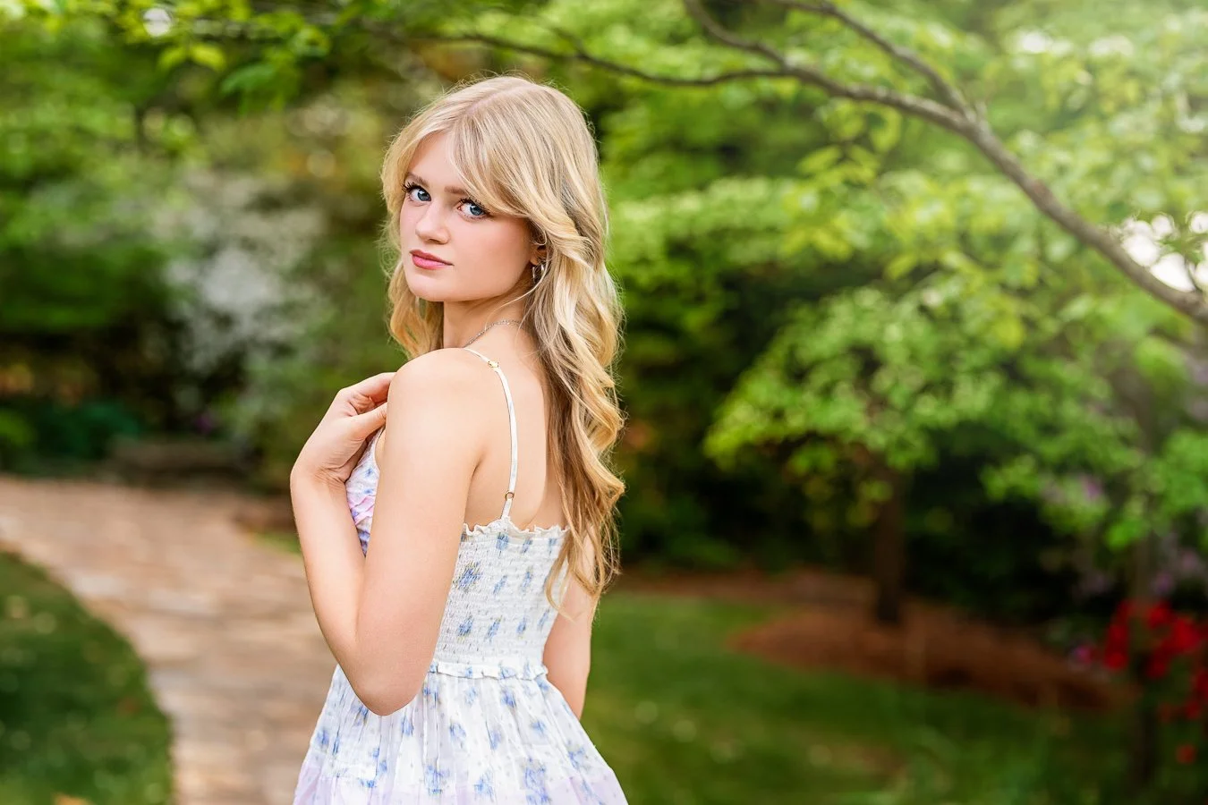 A young blonde woman with blue eyes and light makeup standing outdoors on a garden pathway in Concord NC, surrounded by green trees and bushes, wearing a light-colored floral dress with thin straps.