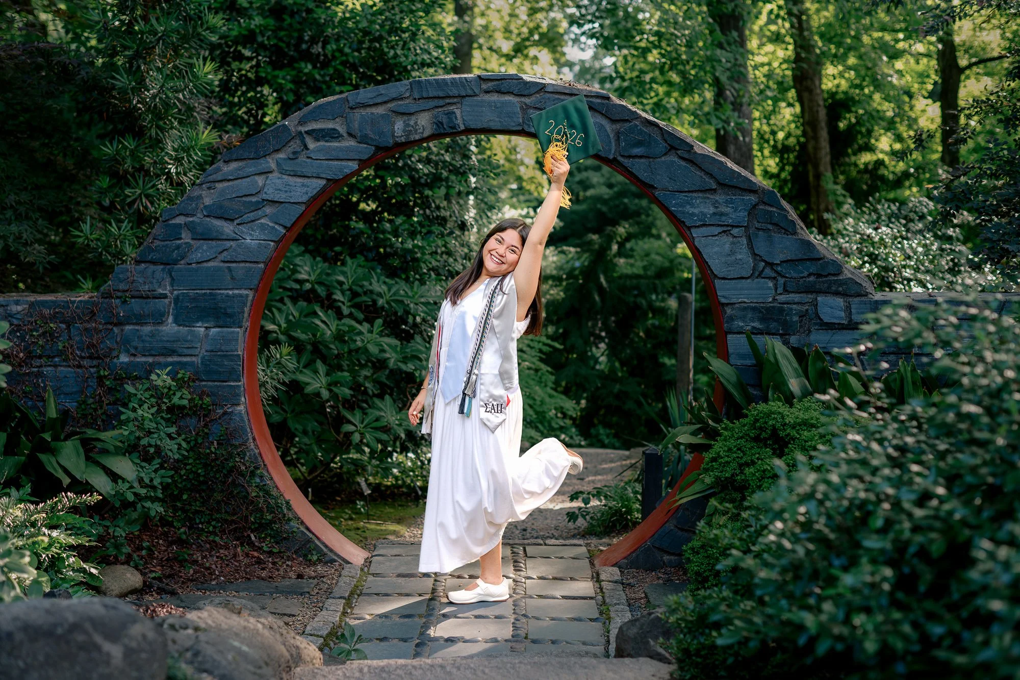 Woman in a white dress and graduation cap kneels on a stone pathway in a lush, green garden at the UNCC botanical gardens, holding a green graduation cap with gold tassels and year '2026' embroidered on it, smiling happily.