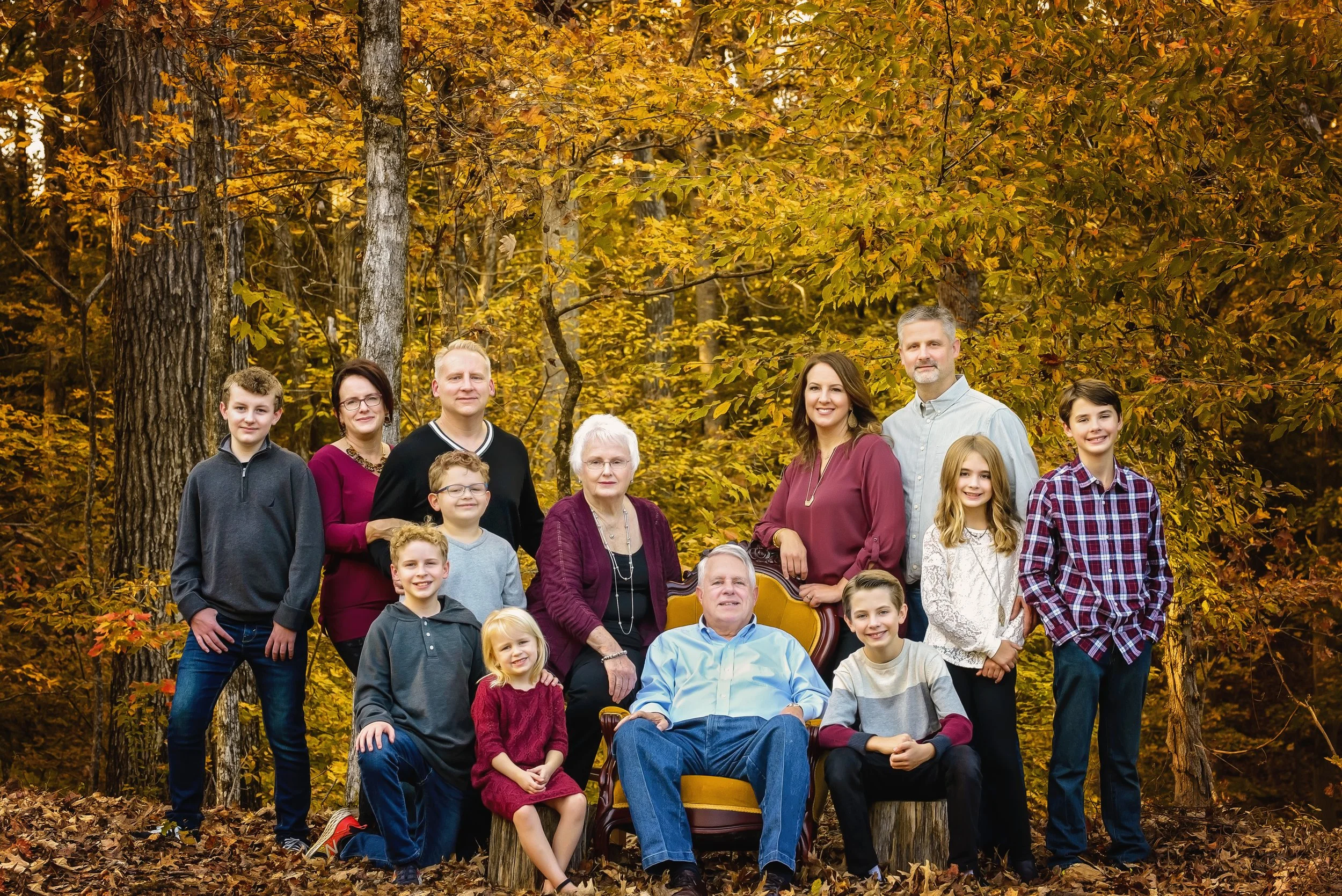 Family portrait of 14 people outdoors in a forest with fall foliage, including grandparent couple, middle-aged parents, teenage, and children.