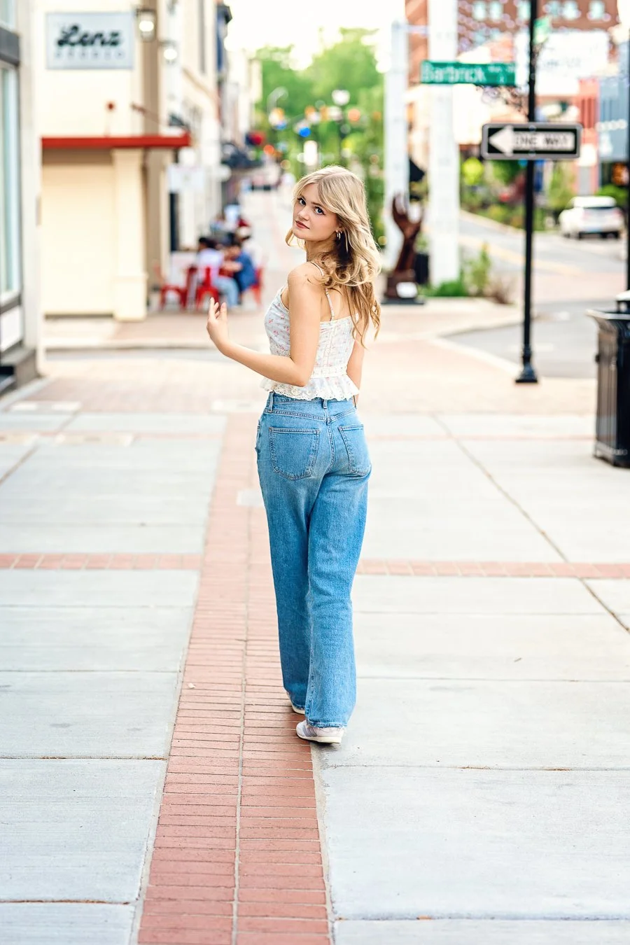 A young woman with blonde hair, wearing a sleeveless white floral top and high-waisted blue jeans, standing on a city sidewalk and looking over her shoulder photographed in Concord NC