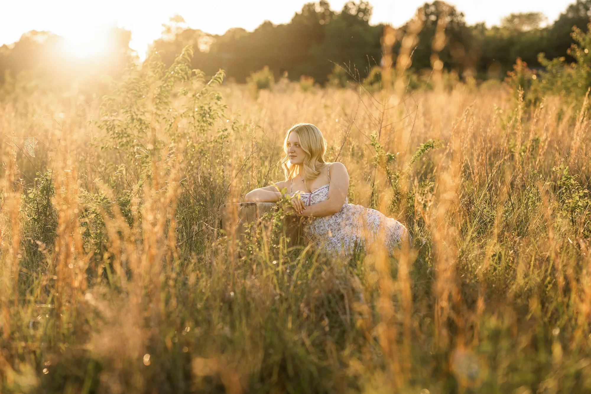 A woman with blonde hair sitting in a field of tall grass and wildflowers at sunset, with sunlight creating a warm glow.