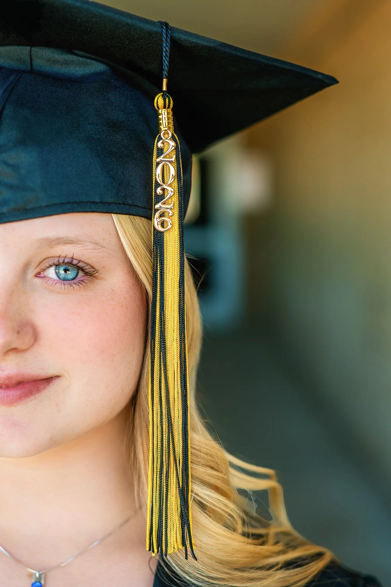 Close-up of a young woman wearing a black graduation cap with a yellow and black tassel that has the year '2020' in silver. She has blonde hair, blue eyes, and is wearing a silver necklace with a small blue pendant, with a blurred green background.