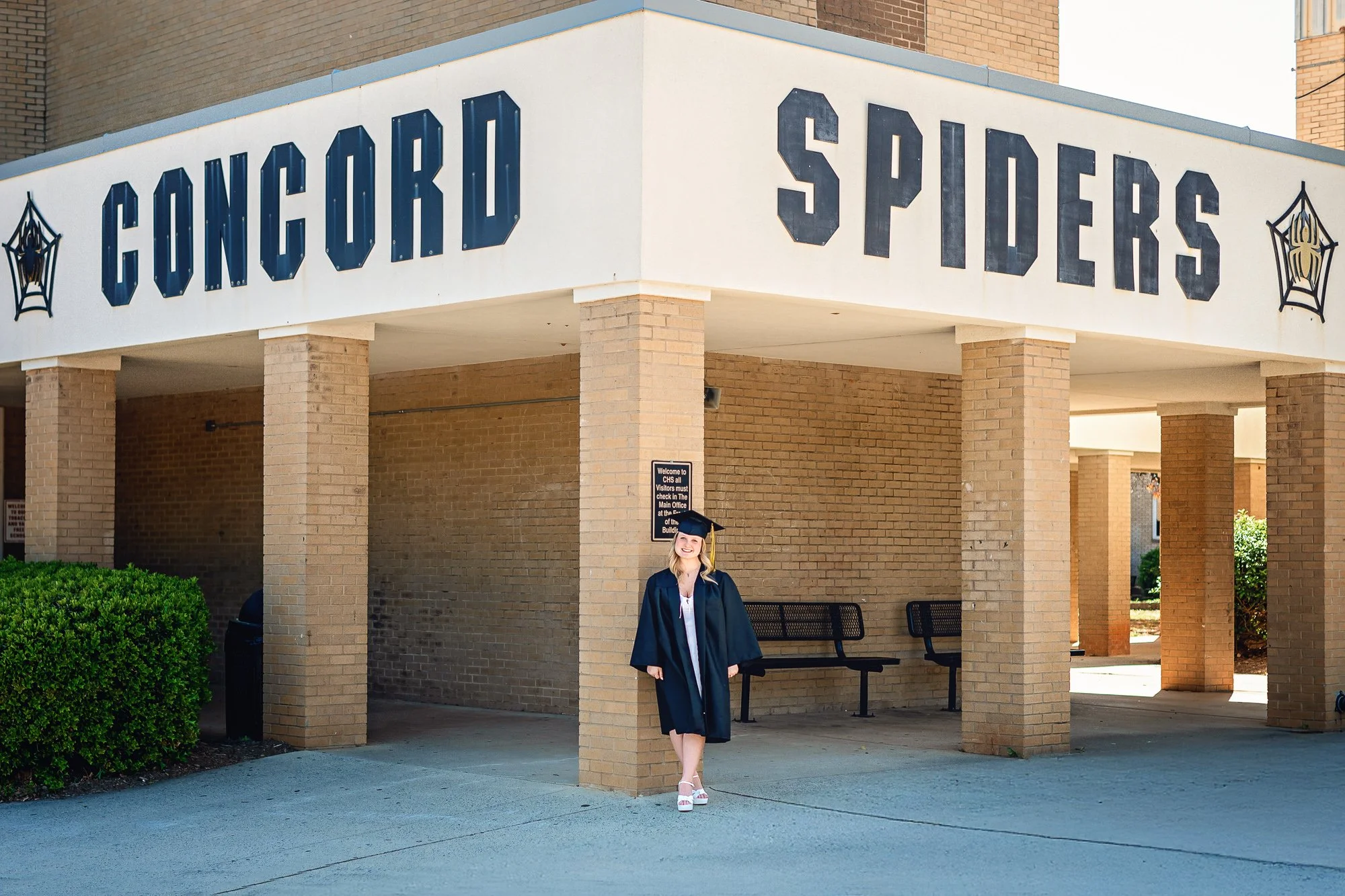 A young woman in a graduation gown and cap standing outside a school building with a sign that reads "Concord Spiders" and spiderweb decorations, celebrating her graduation at Concord Highschool in Concord NC