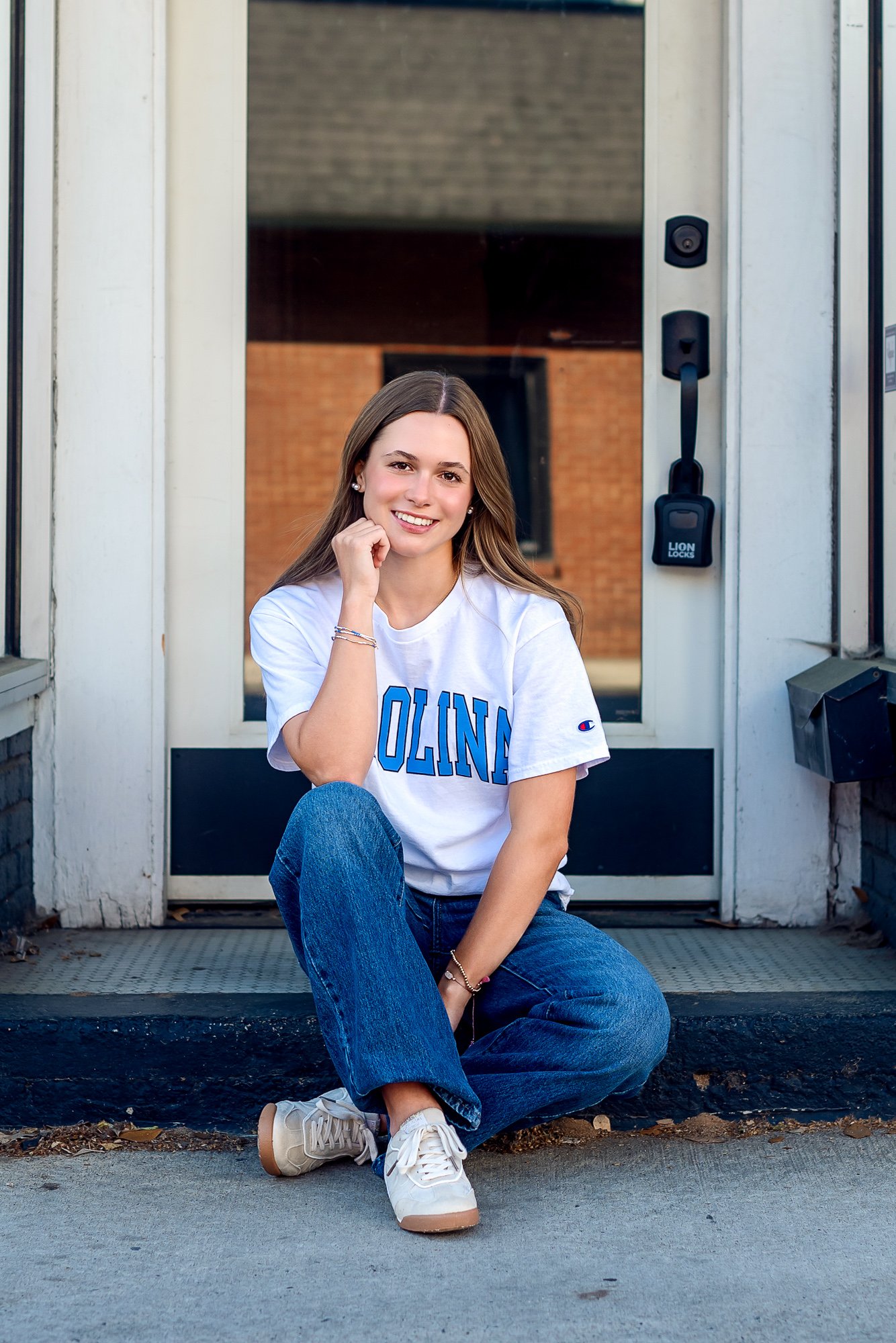 A young woman with long brown hair sitting on the steps in front of a door, smiling at the camera, wearing a white UNC t-shirt with blue lettering, blue jeans, and white sneakers.