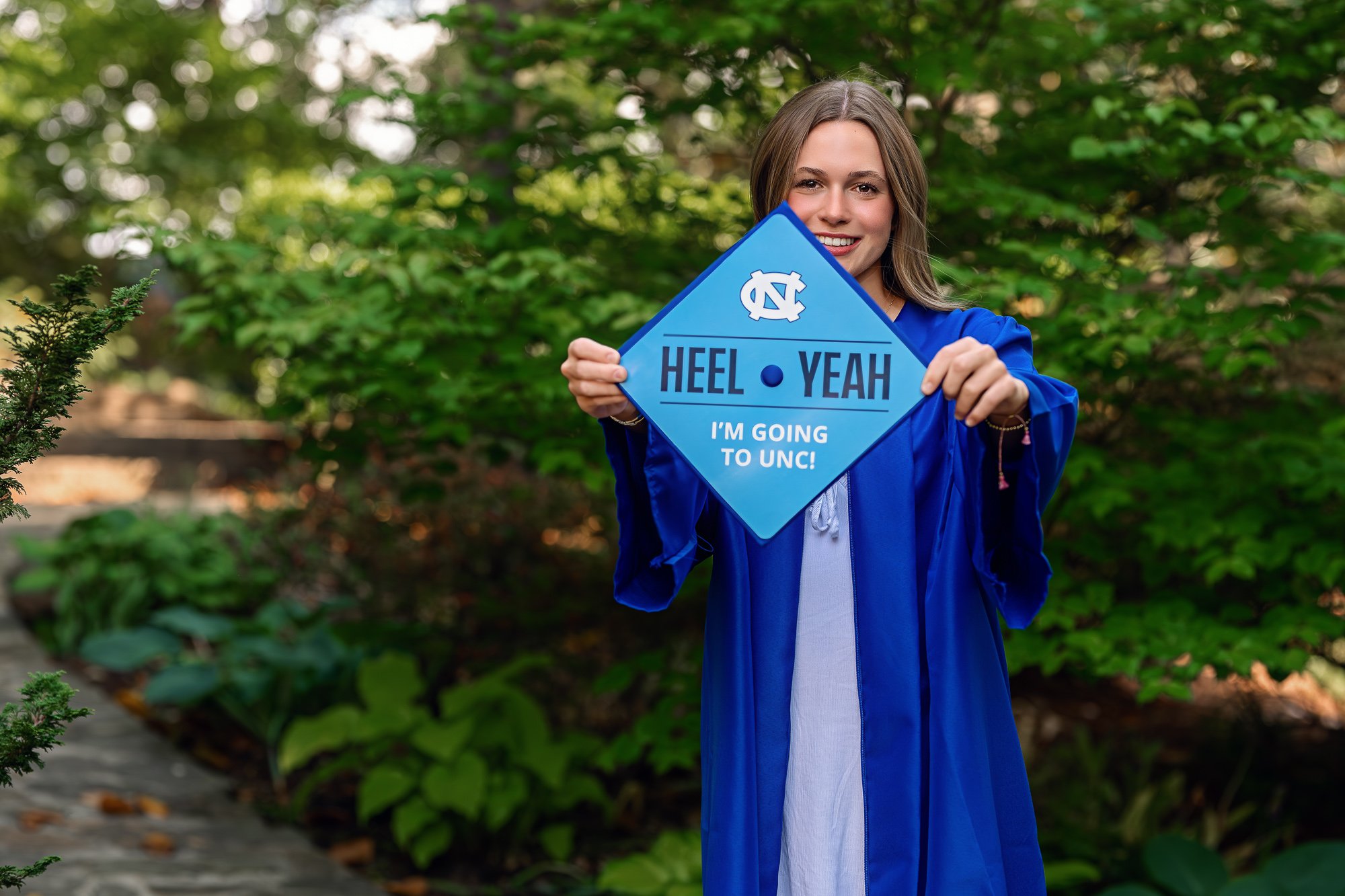 A young woman in a blue graduation gown smiling and holding a blue sign with the University of North Carolina logo, text saying 'Heel Yeah' and 'I'm Going to UNC!' in a lush green outdoor setting in Concord, NC.