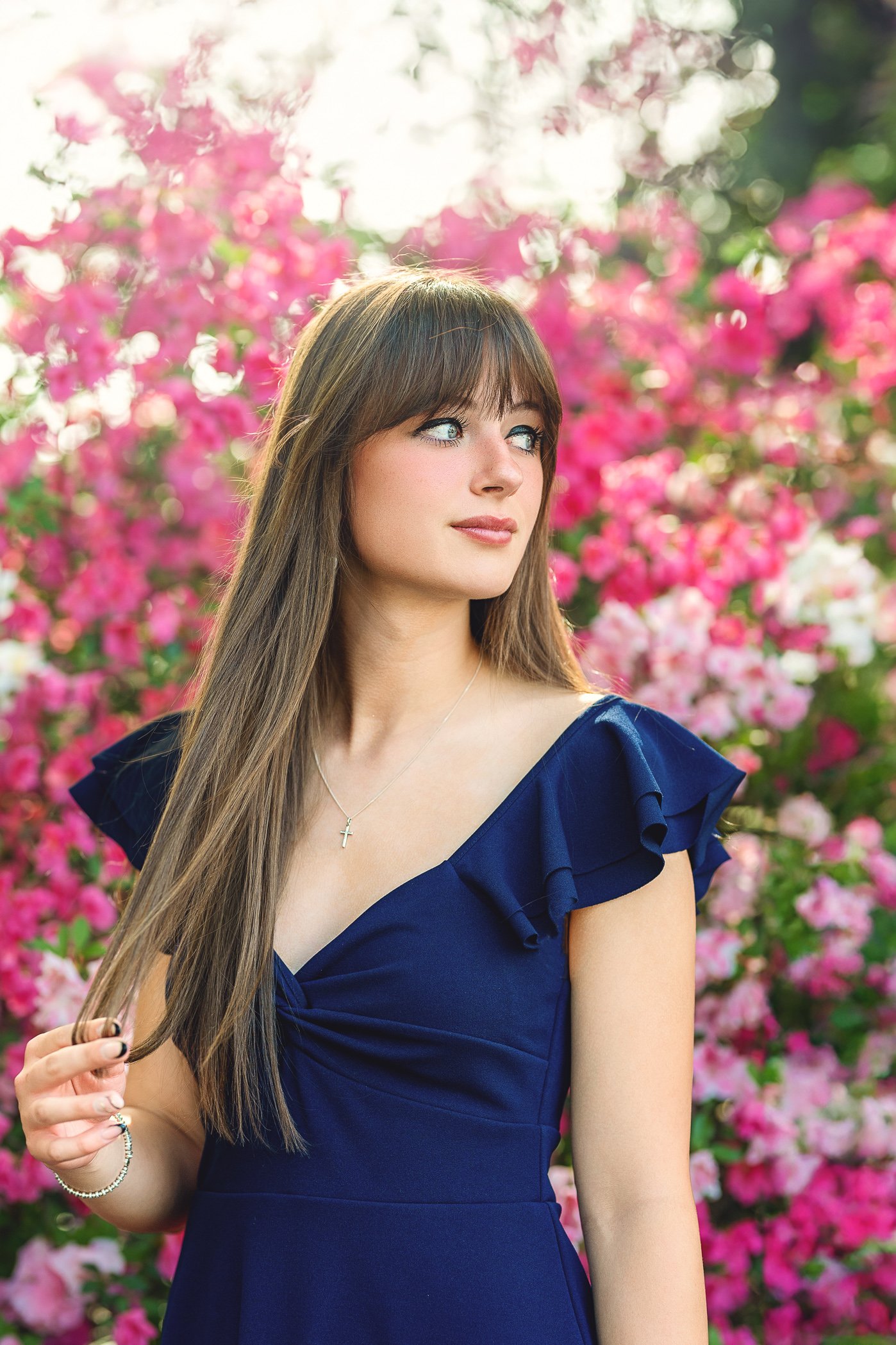 A young woman with long brown hair and blue eyes standing in front of pink flowers, wearing a navy blue dress with ruffled sleeves, a silver cross necklace, and a silver bracelet photographed in the gardens in Concord NC