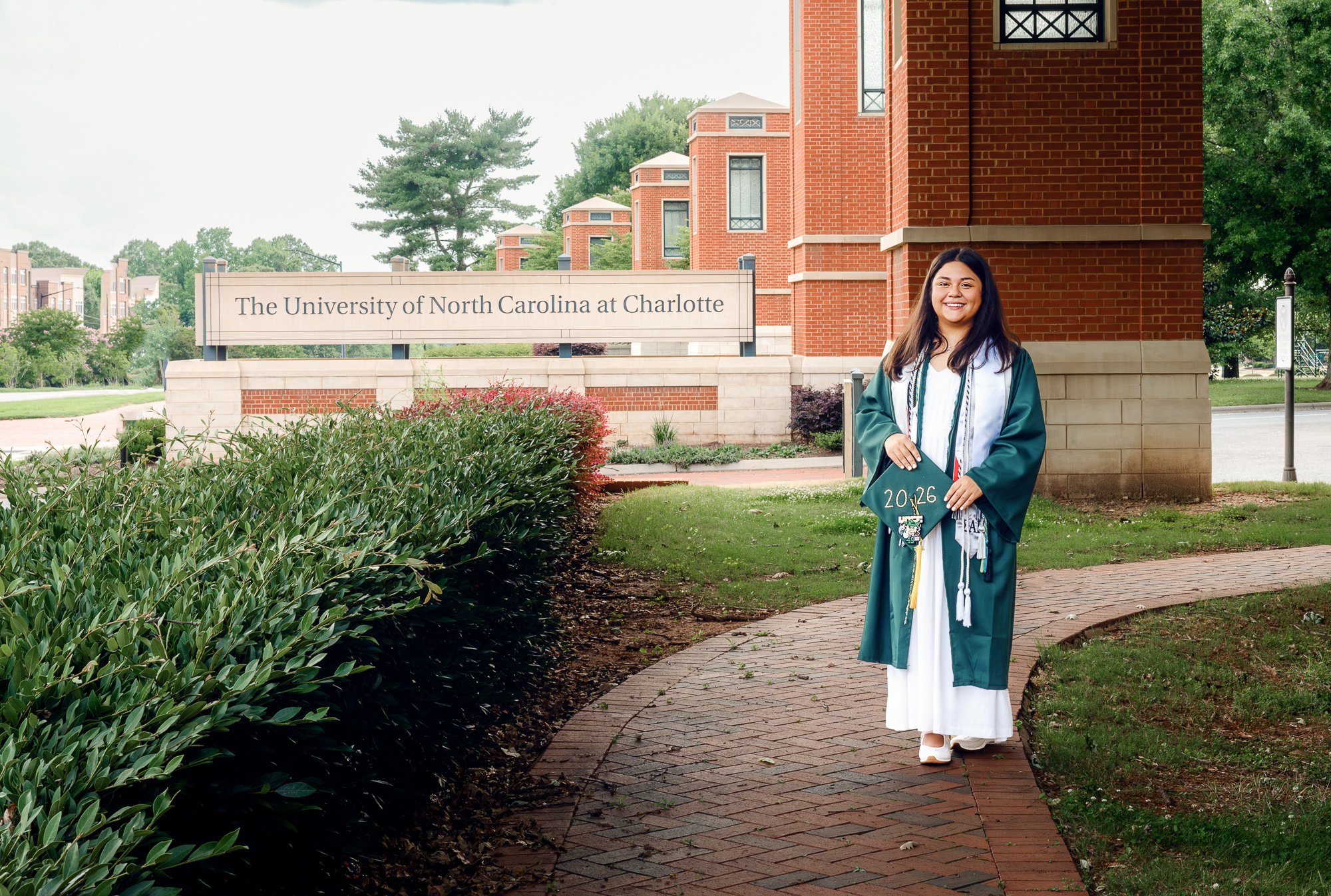 A young woman in a green and white graduation gown and cap standing on a brick pathway in front of a brick building and sign that reads "The University of North Carolina at Charlotte," holding a diploma covered in green fabric with the year 2026 writ