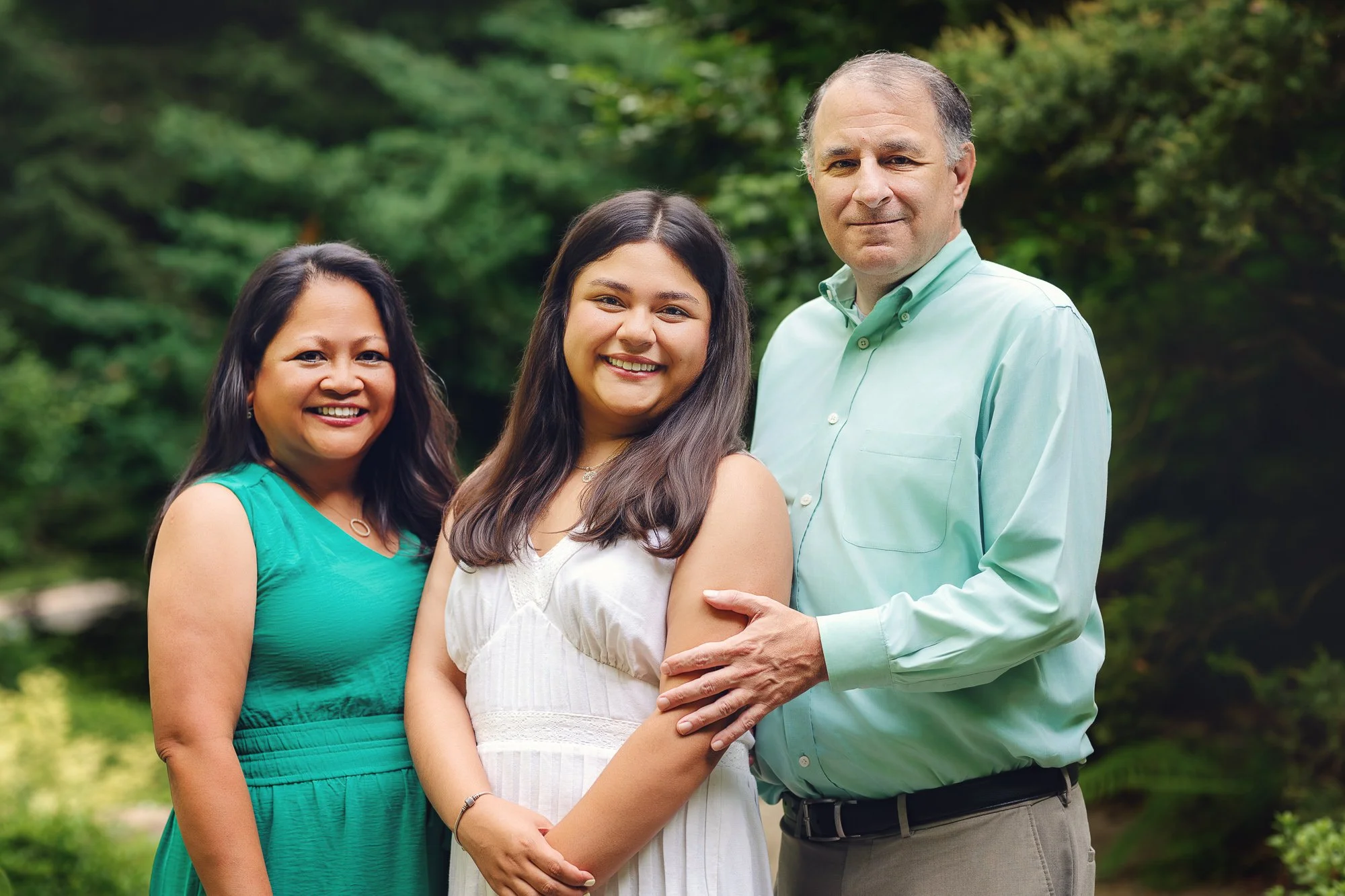Ashley standing between her parents and smiling at the camera in a garden surrounded by green foliage.
