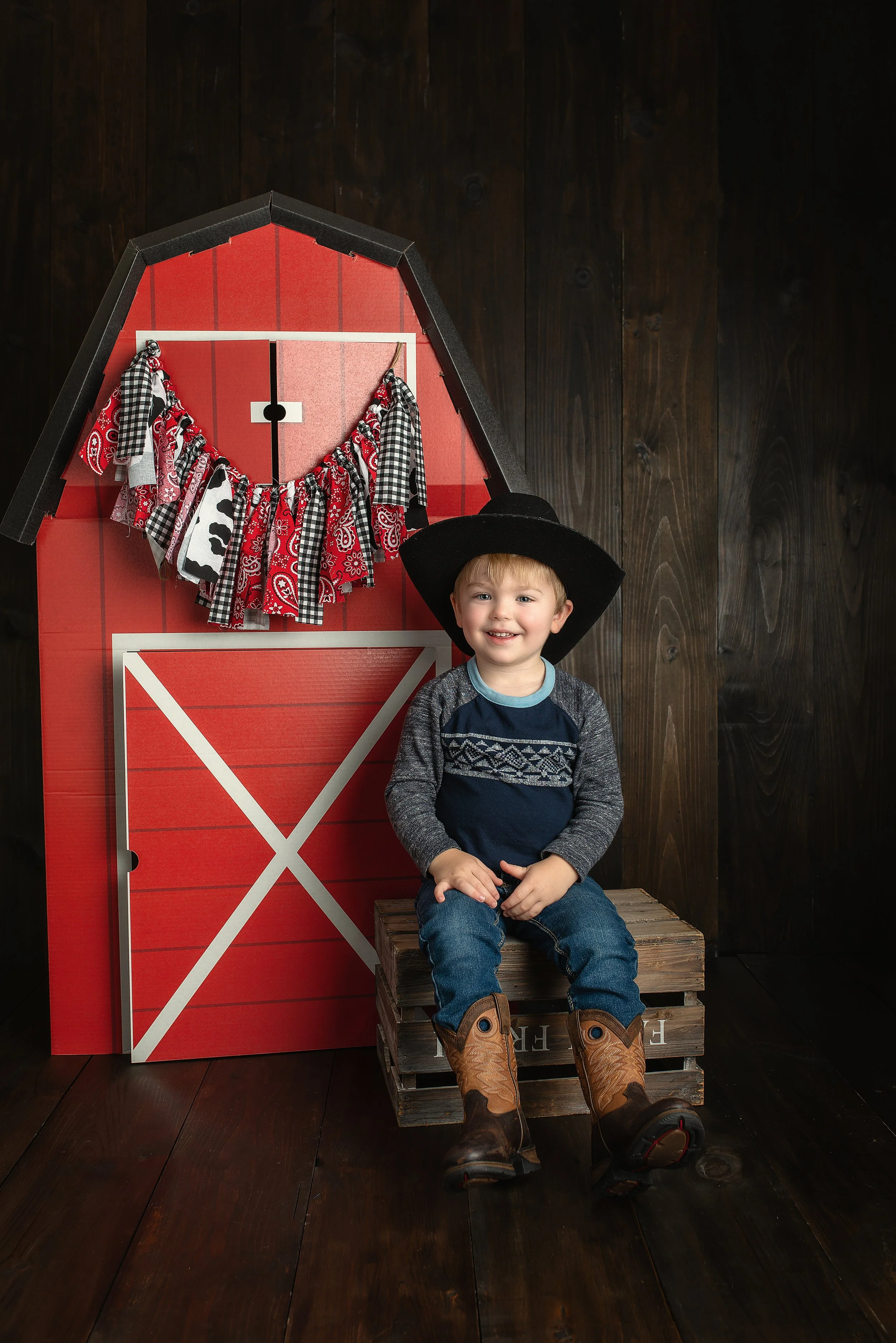 A young boy dressed like a cowboy, sitting on a wooden crate, wearing cowboy boots and a large black hat, smiling in front of a red barn backdrop with a garland of red, black, and white bandana-patterned fabric.
