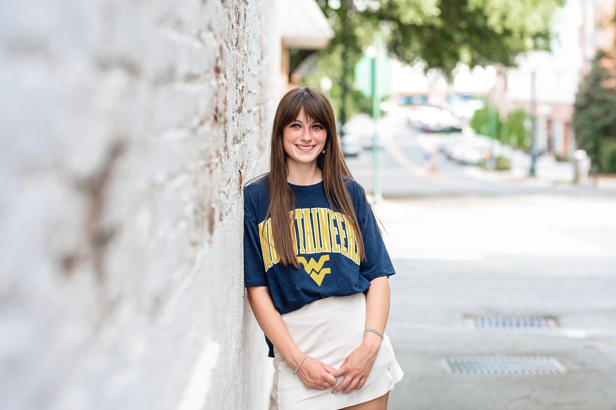 A young woman with brown hair and blue eyes smiling while leaning against a white brick wall outdoors. She is wearing a navy blue t-shirt with yellow and white text that says 'MOUNTAIN W.VA.' and a white skirt. The background shows a city street with