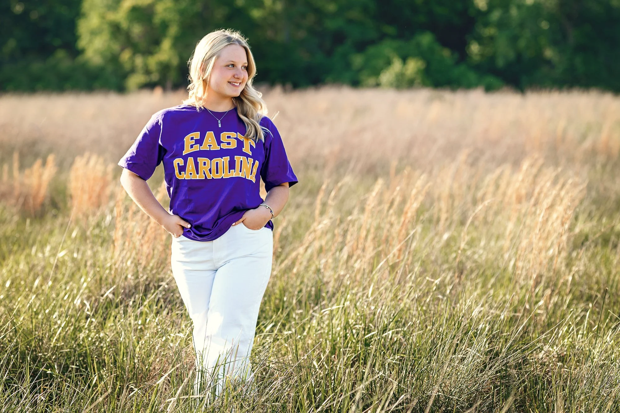A young woman with blonde hair wearing a purple 'East Carolina' T-shirt and white pants standing in a grassy field with a background of trees, smiling and looking to her right.