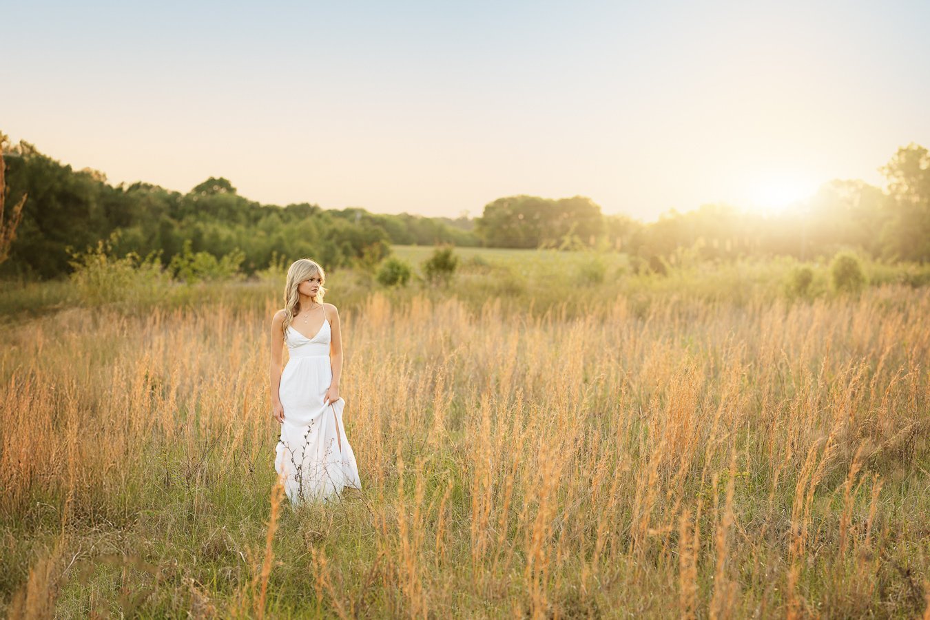 A woman in a white dress standing in a field of tall grass as the sun sets on the horizon in Harrisburg NC