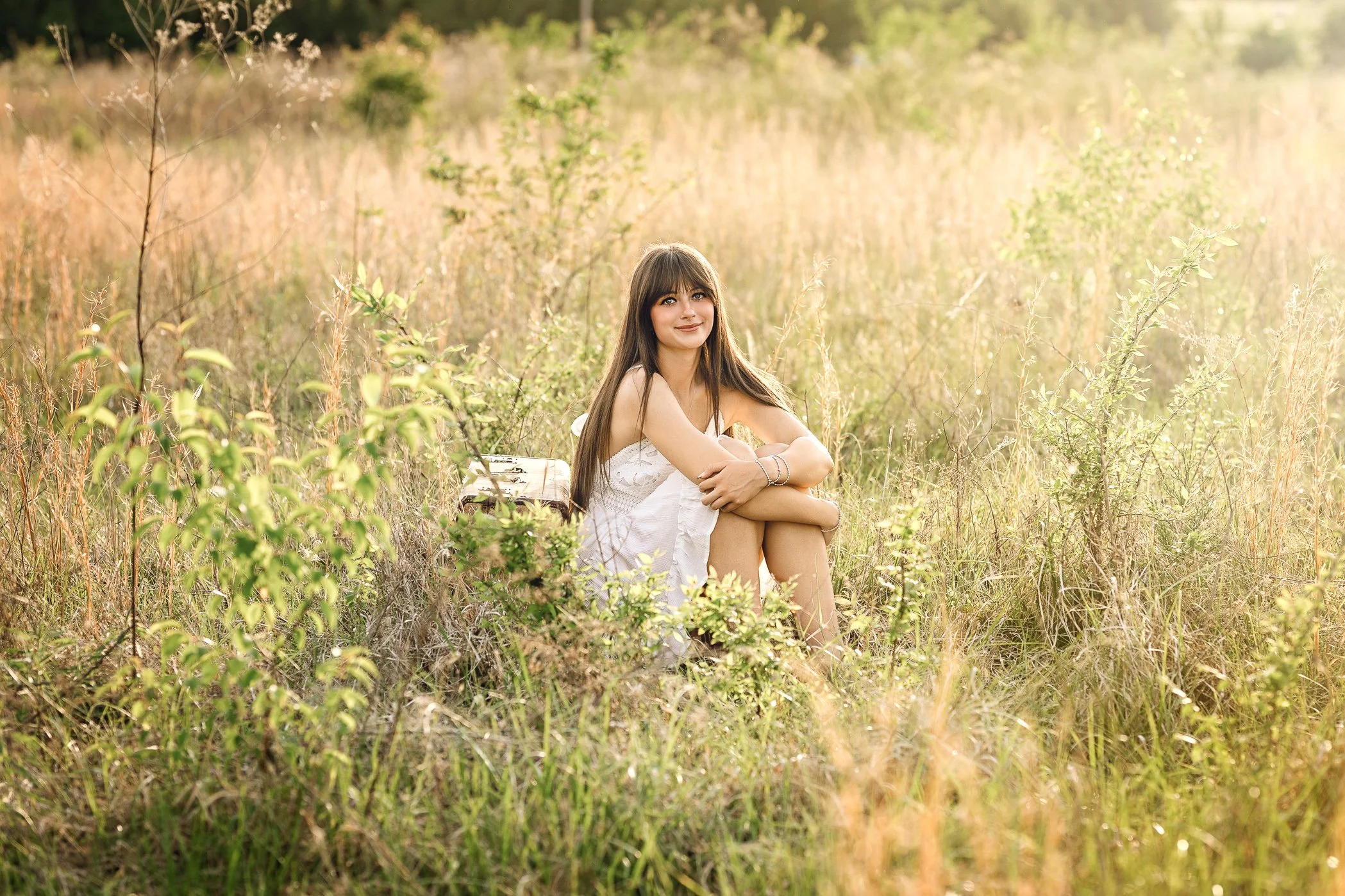 cox mill high school senior girl in white dress with long dark hair and bangs photographed in open grassy field with bright sunlight. She's seated in the grass leaning against an old suitcase