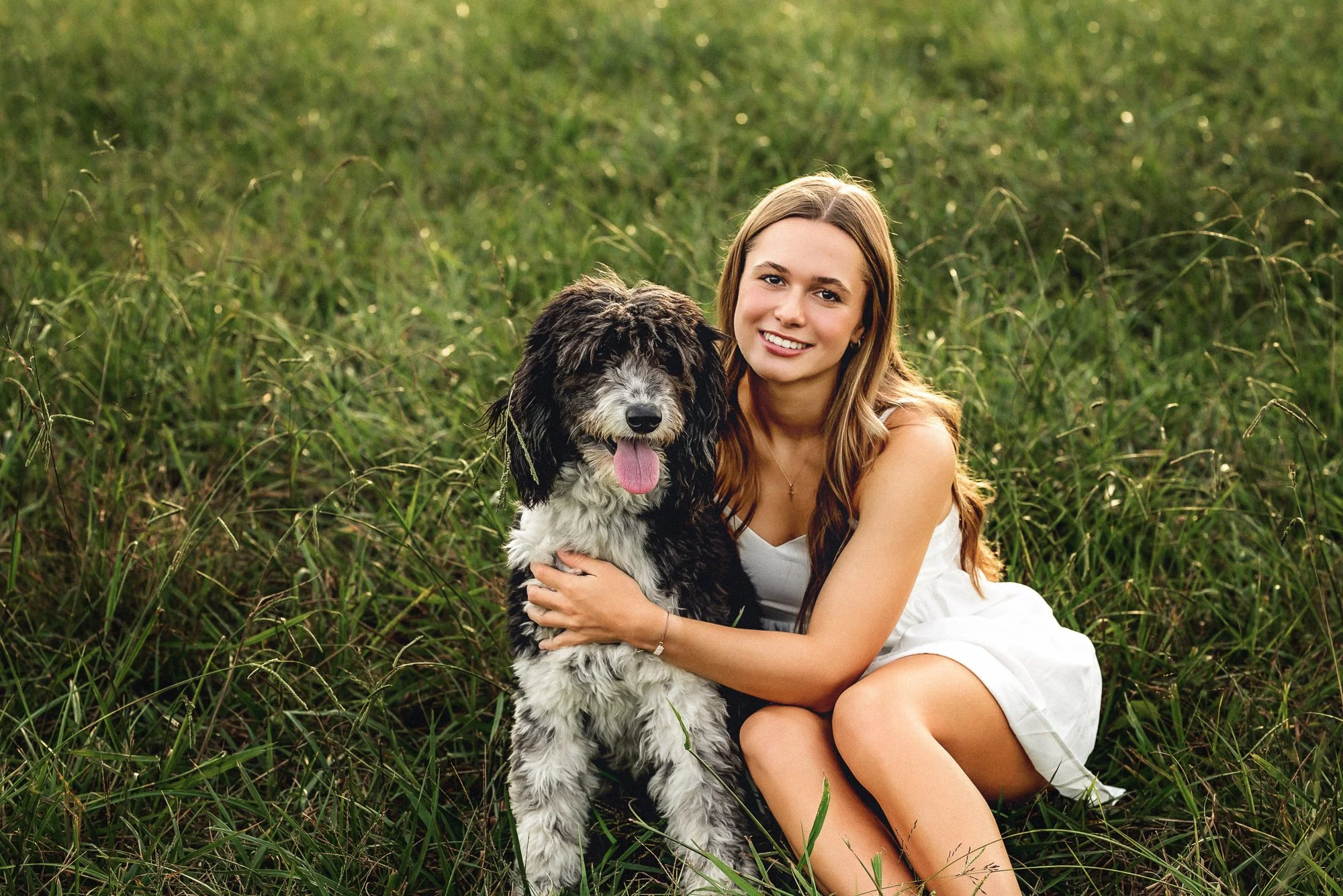 High school senior sitting in a grassy field with her black and white dog during golden hour