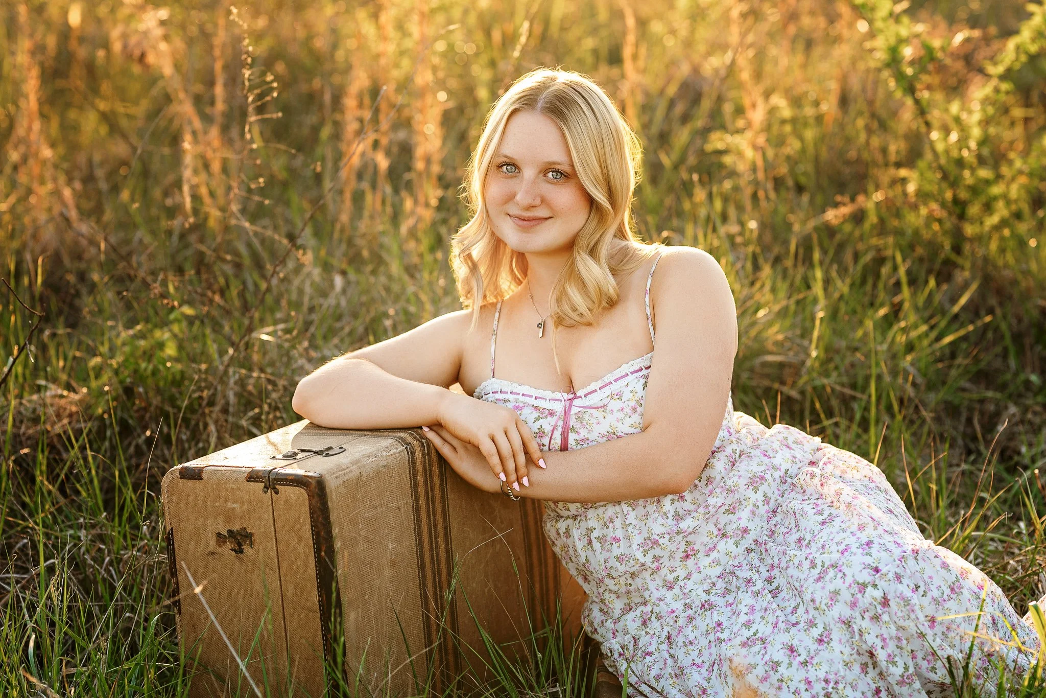 Concord High School Senior girl photographed sitting field leaning against vintage suitcase with golden light behind her