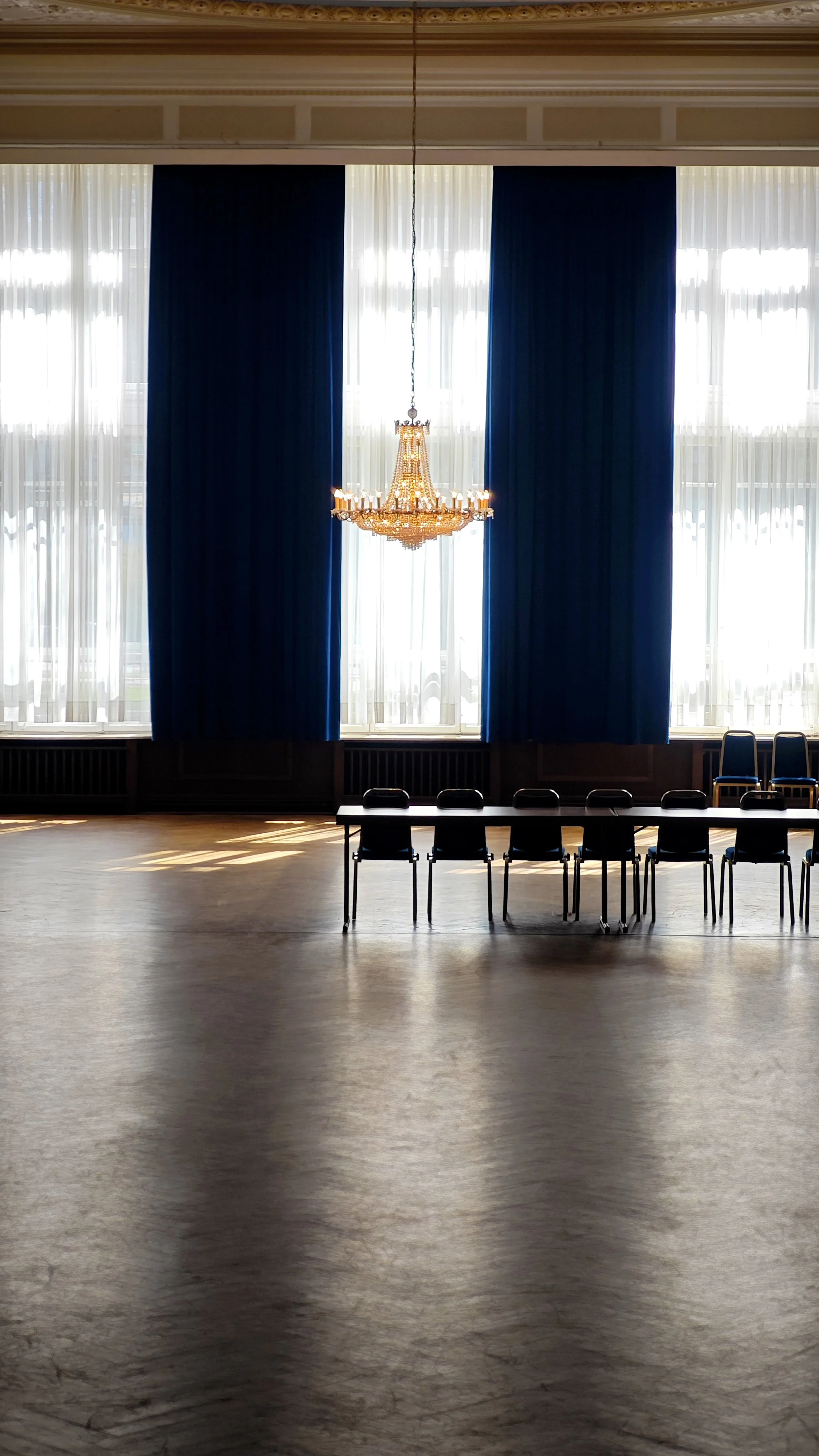 Ein Saal mit großen Fenstern, blauen Vorhängen, einem Kronleuchter in der Mitte und mehreren Stühlen.