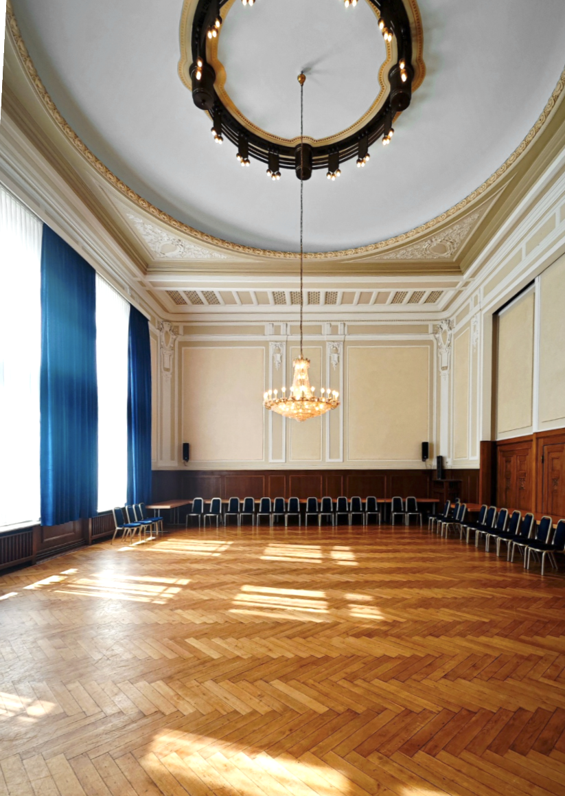 Ein großes, elegant gestalteter Saal mit hölzernem Parkett, hohen Fenstern mit blauen Vorhängen, einer prächtigen Decke und einem großen Kronleuchter in der Mitte.