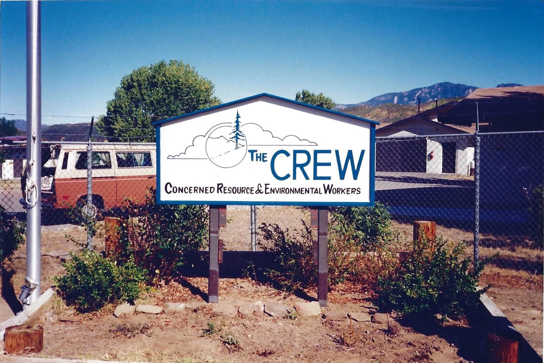 A sign reading 'The CREW Concerned Resource and Environmental Workers' in front of a chain-link fence, with a pickup truck and a mountain range in the background.