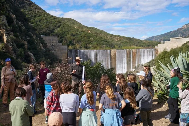 Tour group listening to guide near a dam and waterfall, surrounded by mountains and desert vegetation.