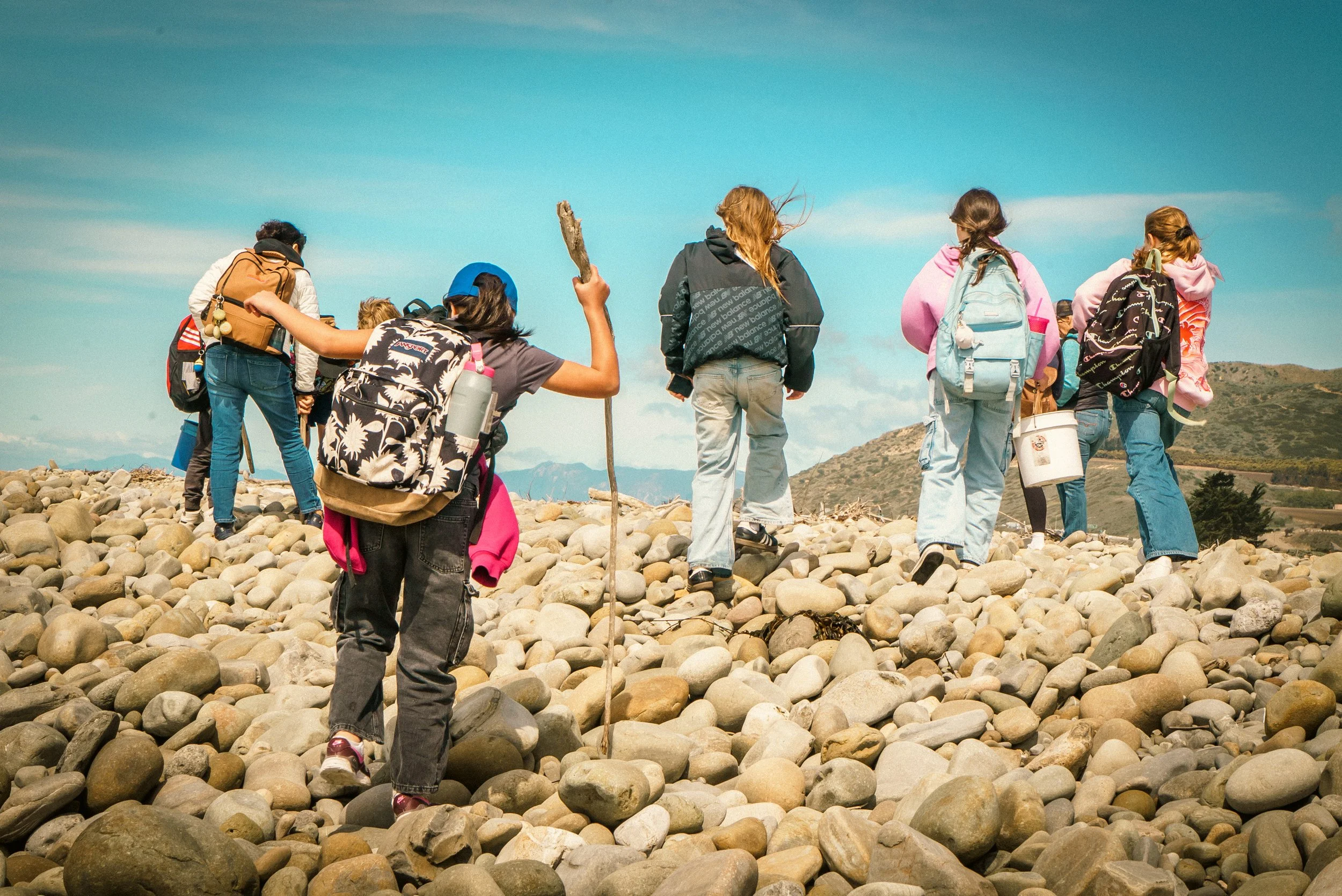 Group of children walking on a rocky beach, carrying backpacks and sticks, with mountainous terrain and blue sky in the background.