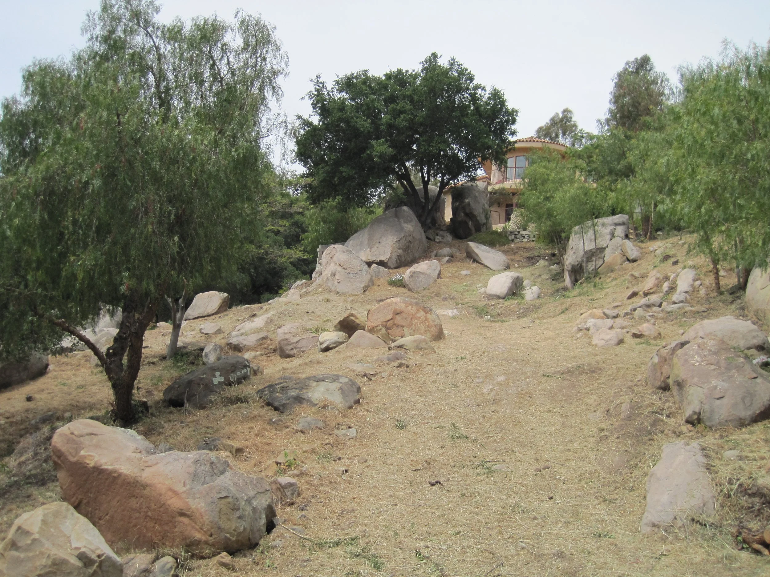 A dirt area with scattered large rocks and trees in front of a house on a hill, with a cloudy sky.