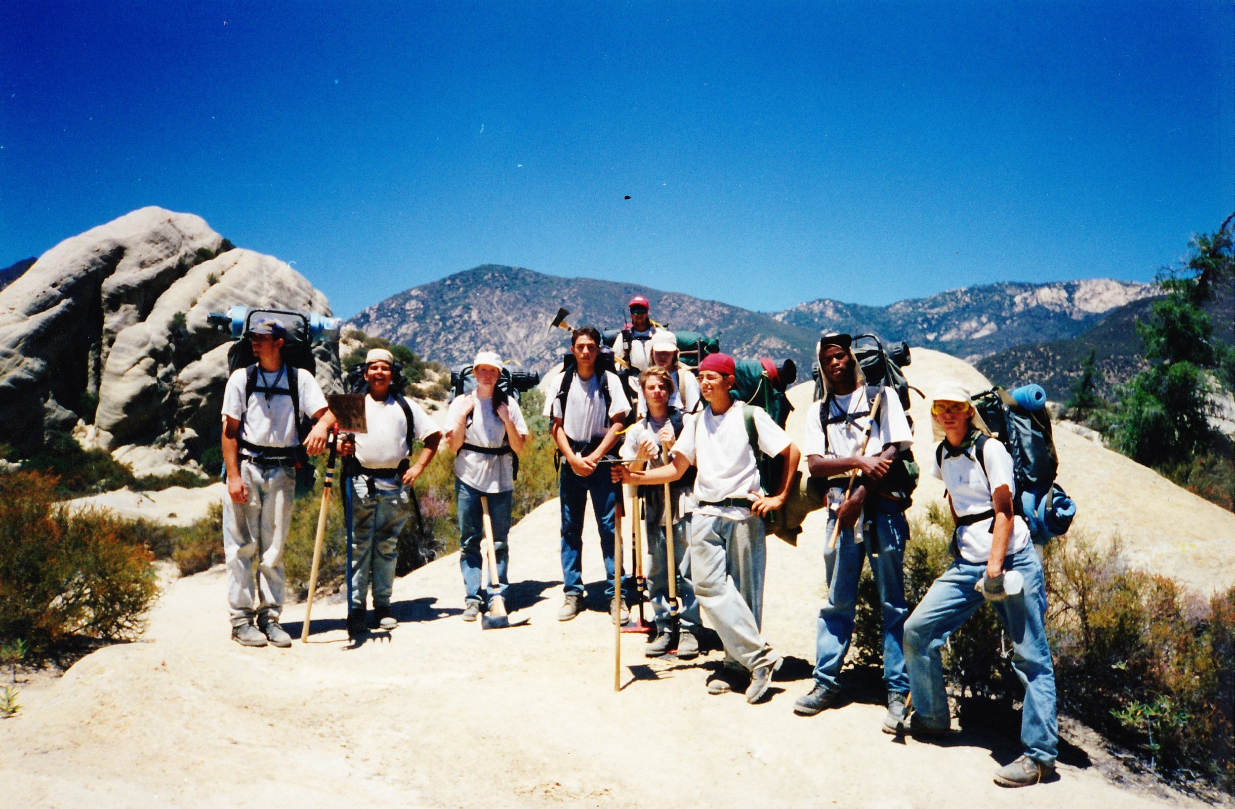 A group of people dressed in hiking gear standing on a trail in a mountainous area with large rocks, trees, and distant mountain ranges under a clear blue sky.