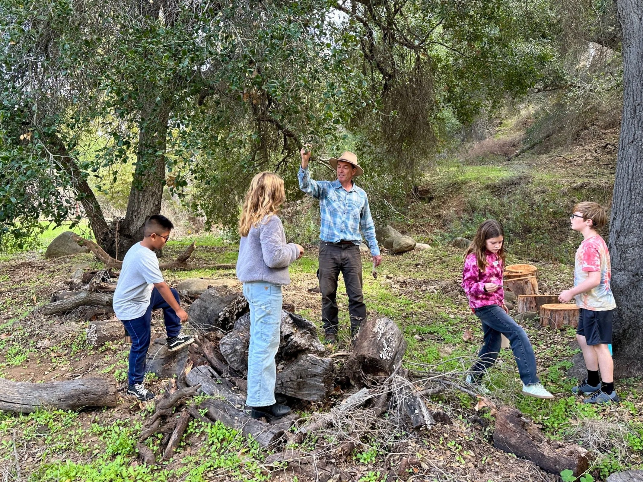 A group of children and an adult in a wooded outdoor area, with a man in a blue plaid shirt and a wide-brimmed hat showing them something. The children are standing around and looking at what the adult is indicating, with some sitting or standing among logs and rocks.