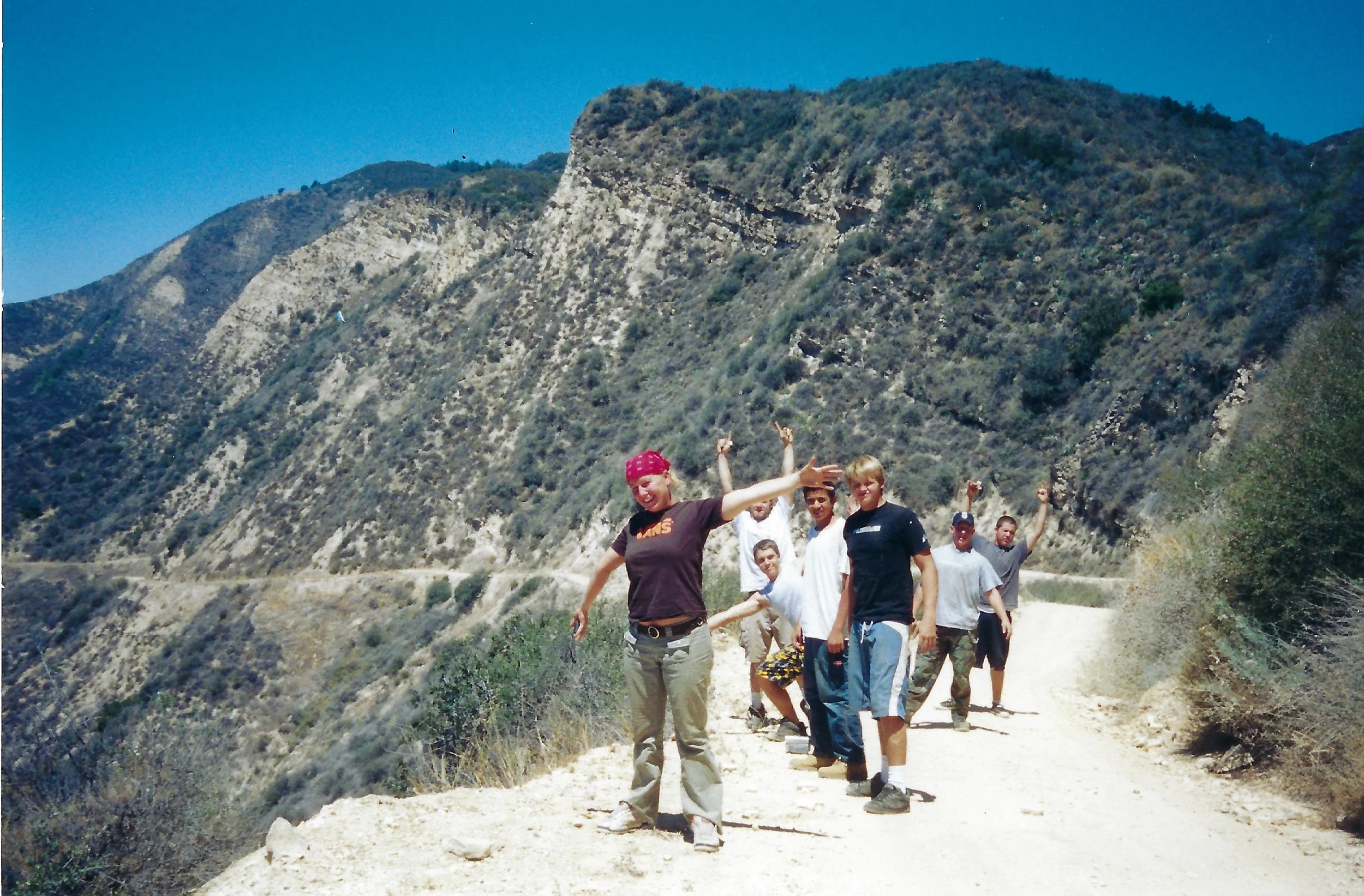 A group of people hiking on a dirt trail in a mountainous area with dry, rugged terrain and trees on the mountain slopes.