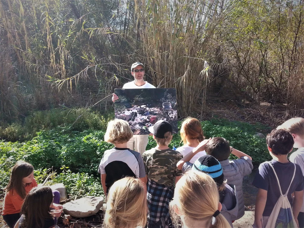 A man holds up a photograph for a class of students in front of a stand of giant reed in the Ventura River bottom