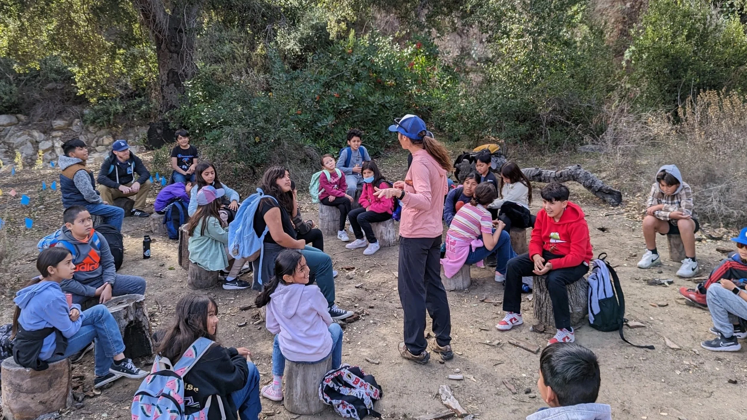 A group of children sitting outdoors in a forested area, listening to a woman standing and talking to them during a field trip or outdoor class.