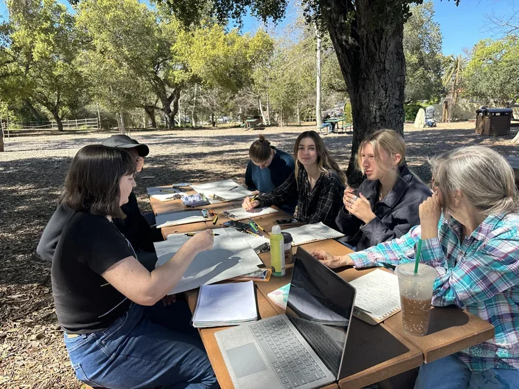 A group of six women sitting at a wooden table outdoors under a large tree, engaged in a meeting or discussion with notebooks, laptops, and drinks.