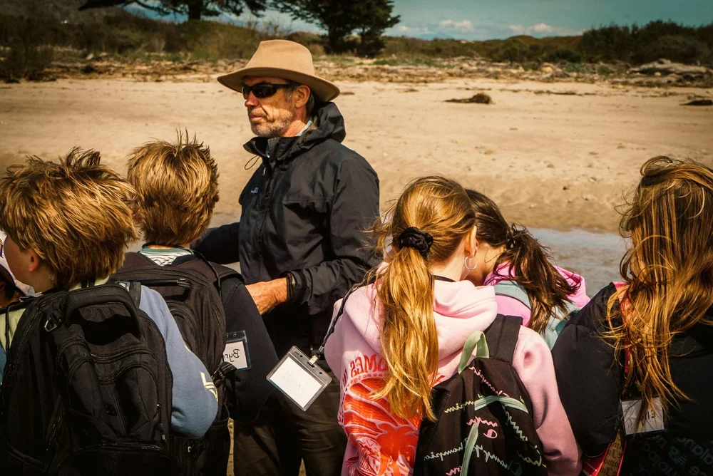 A man in a wide-brimmed hat talks with students wearing backpacks on the sand at the estuary
