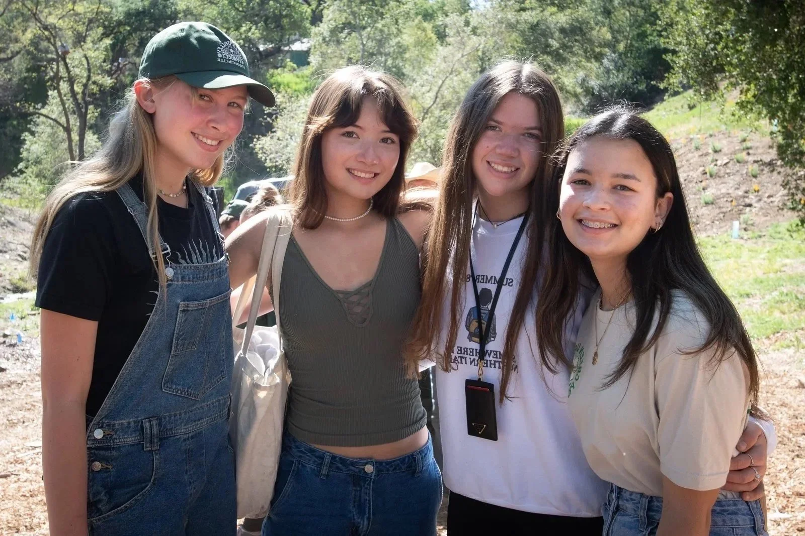 Four young women standing outdoors on a sunny day, smiling at the camera, with trees and grassy area in the background.