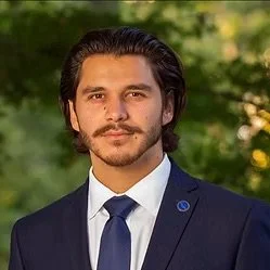 A man with dark hair, a beard, and mustache, wearing a navy suit, white shirt, and blue tie, standing outdoors with blurred greenery in the background.